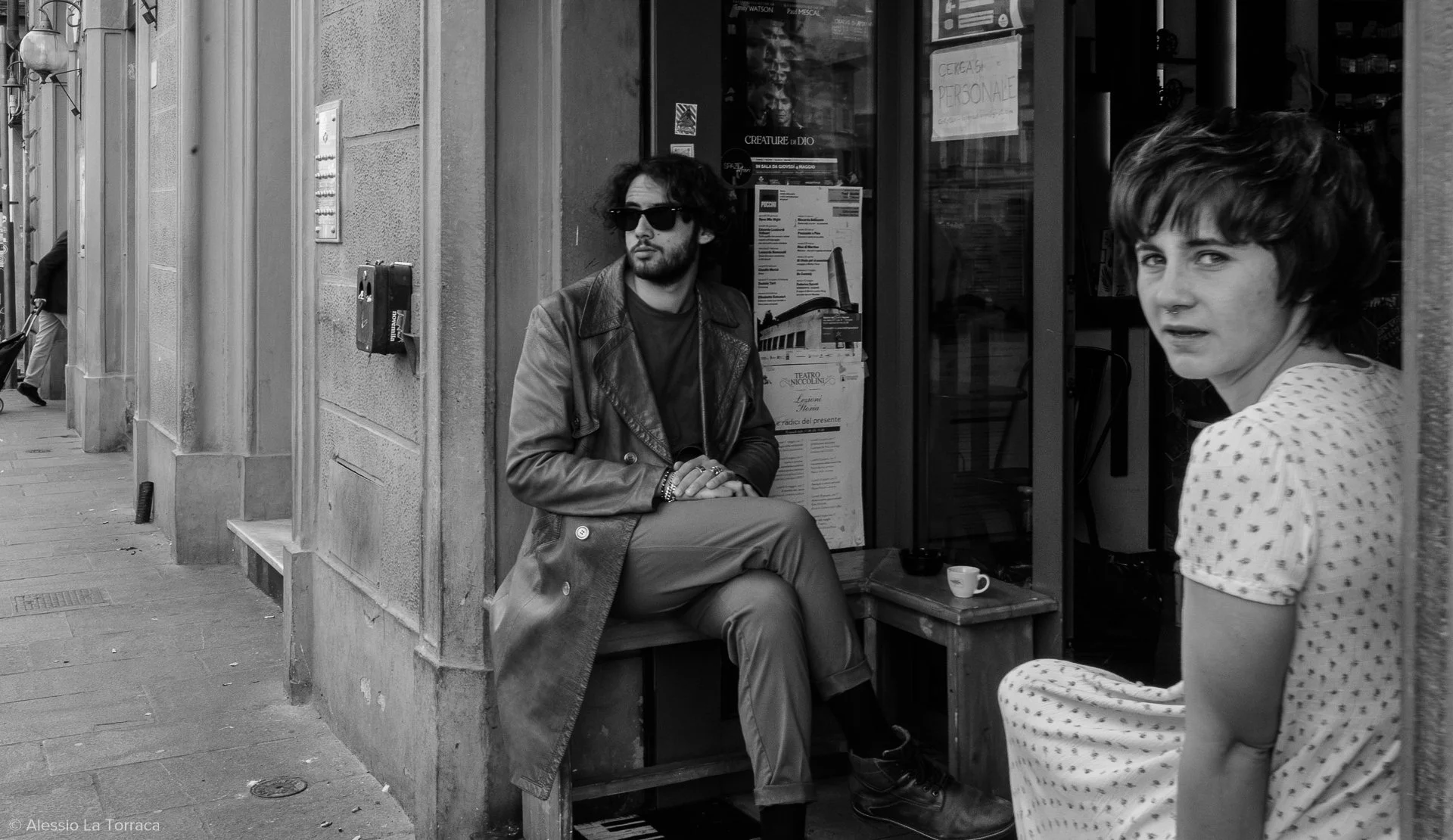 A man with dark curly hair, sunglasses, and a leather jacket sitting on a bench outside a cafe, while a woman with short curly hair and a polka dot dress looks towards the camera. The scene is on a city sidewalk.