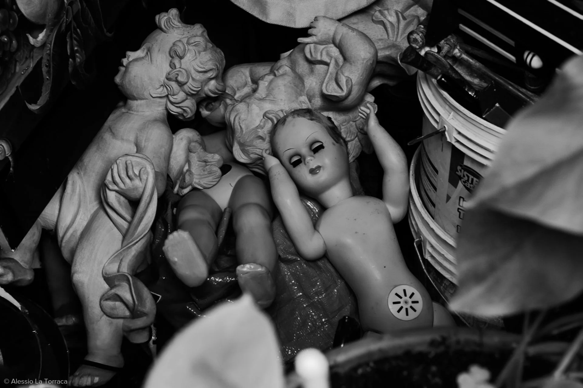 A collection of old, damaged dolls and sculptures, including a broken doll with missing eyes and a painted face, among various other figurines and objects in a cluttered space.