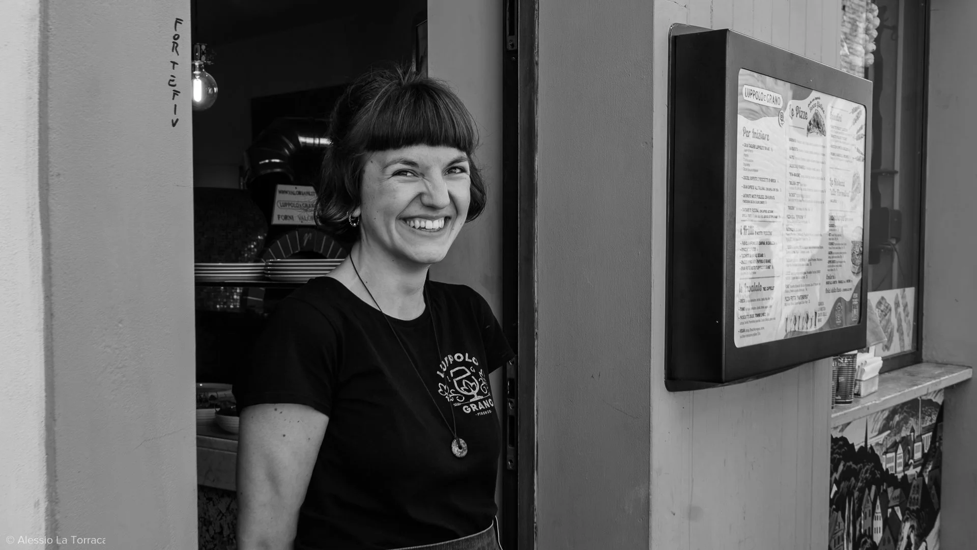 A woman with short hair smiling and standing in front of a restaurant menu display outside. She is wearing a black t-shirt and a necklace.
