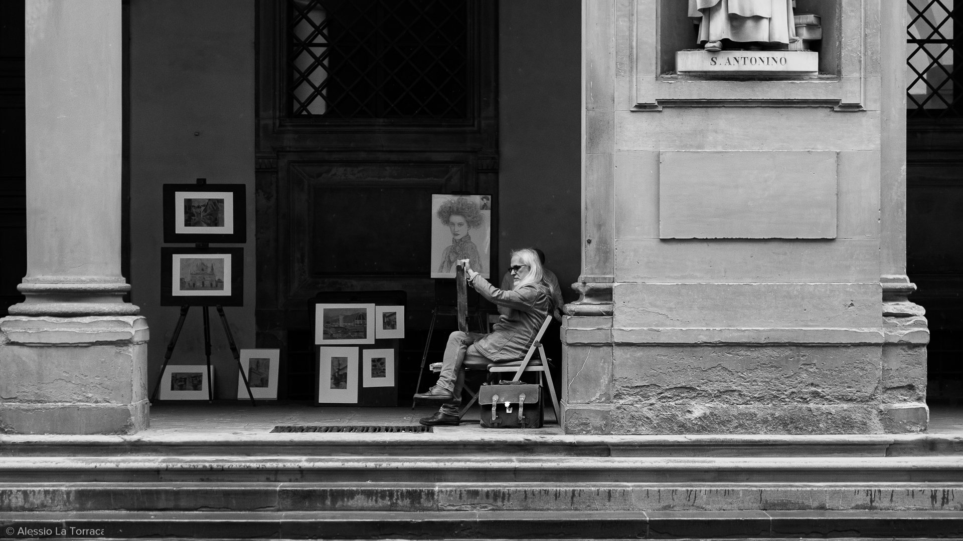 Black and white photo of a female artist sitting on a chair, painting on a canvas with a cityscape. Her art supplies are on a bag nearby, and her artwork includes a portrait of a woman with curly hair. Artworks are displayed on easels and the wall next to her.