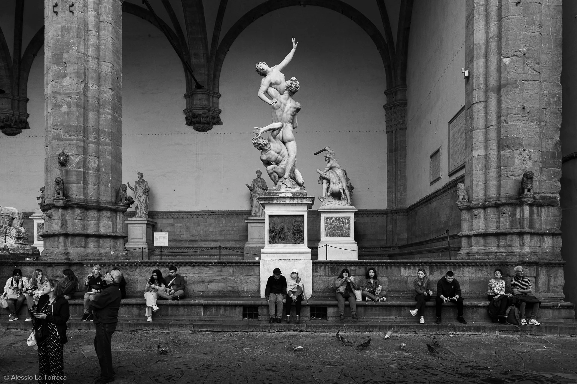People sitting and standing along a stone bench beneath a large classical sculpture in a gallery with stone columns and arches, pigeons on the ground.