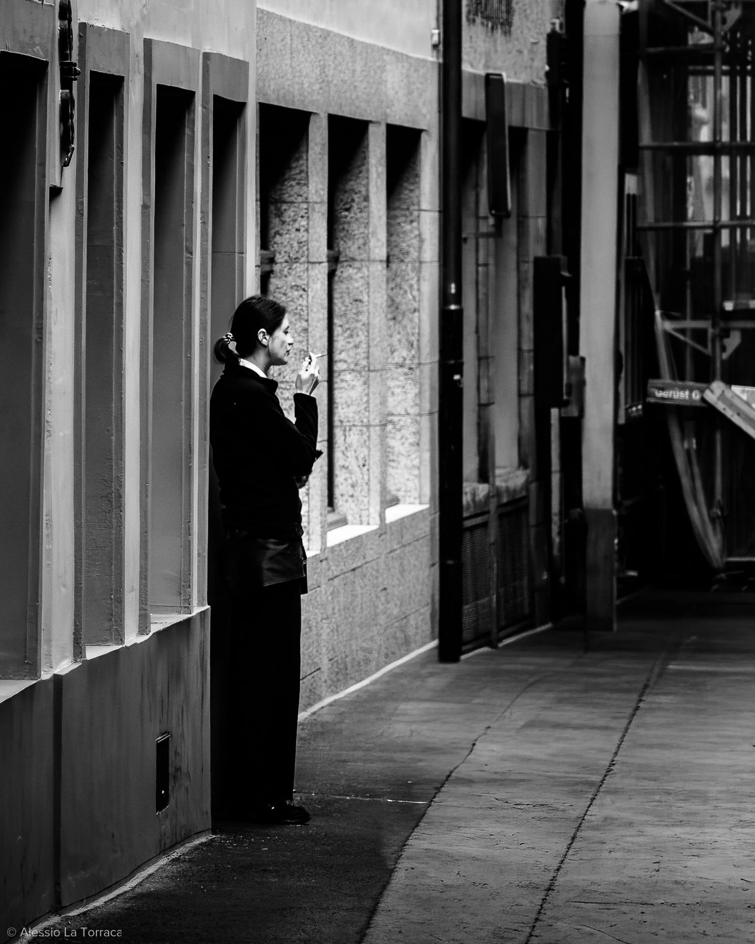 A woman standing on a city sidewalk, leaning against a wall, smoking a cigarette, with construction scaffolding in the background.