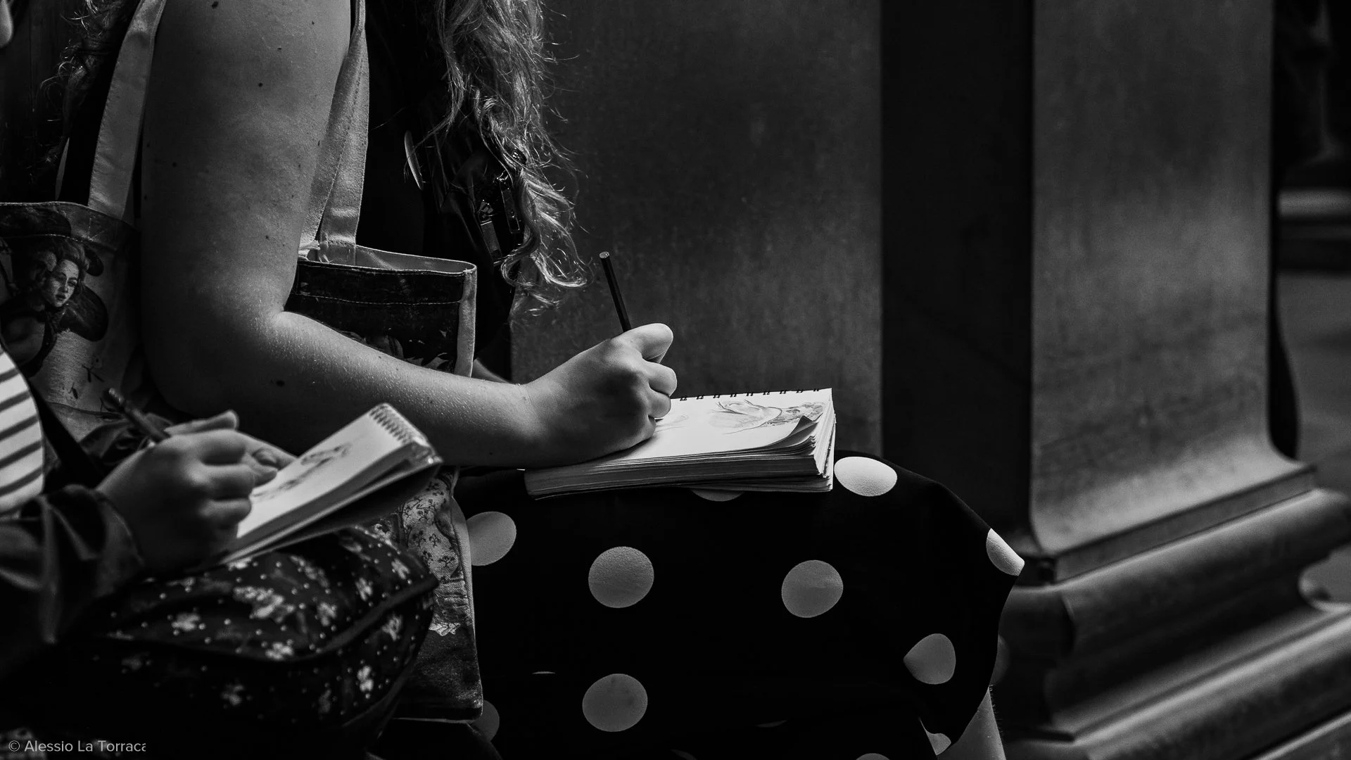 A black and white photo of a person sitting on a bench, sketching in a notebook, with another person's hand holding a small sketchpad nearby.