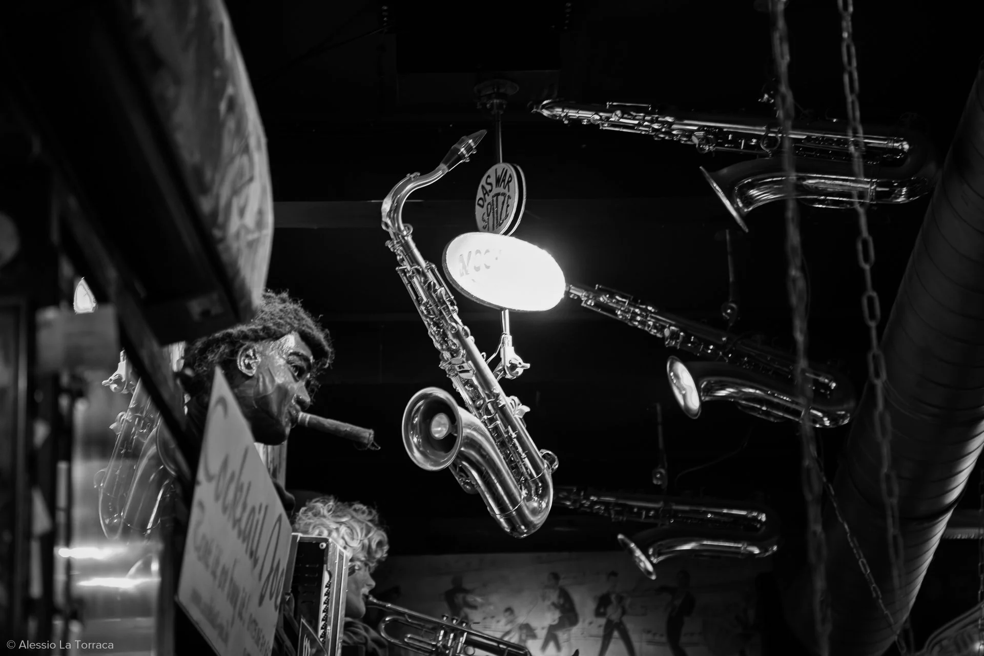 Black and white photo of two jazz musicians playing saxophones in a dimly lit music venue. The musicians are focused, with one smoking a cigar. There are chains hanging from the ceiling and a sign that reads 'DAS WAR ETC' in the background.