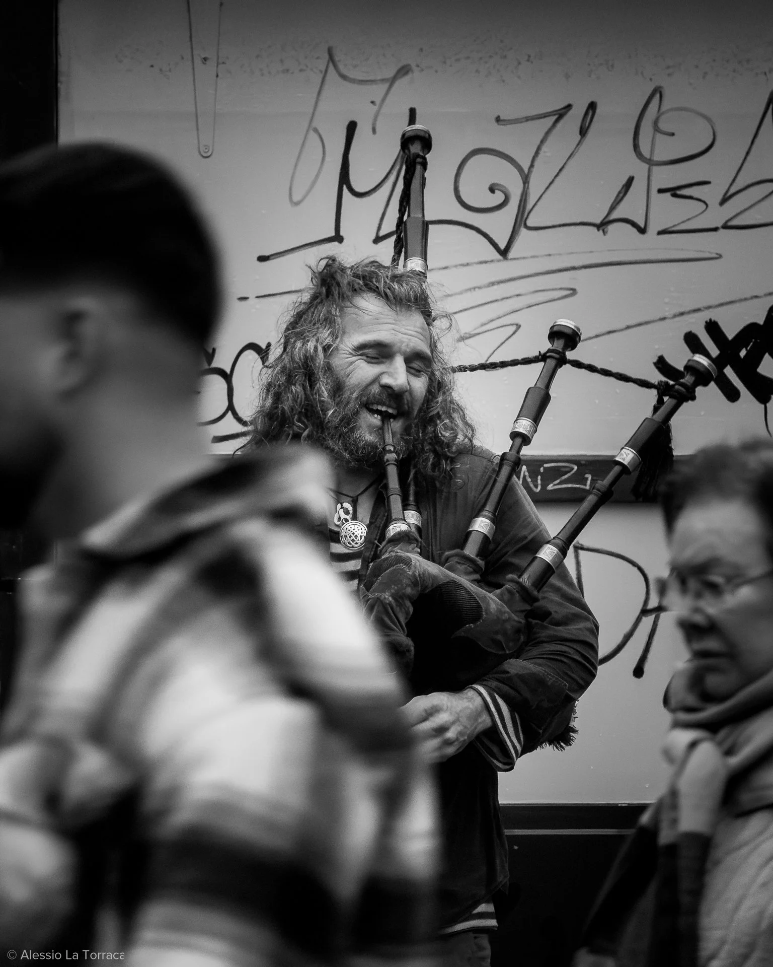 A man with long curly hair and a beard playing bagpipes, surrounded by blurred people in the foreground, with a graffiti-covered wall in the background.