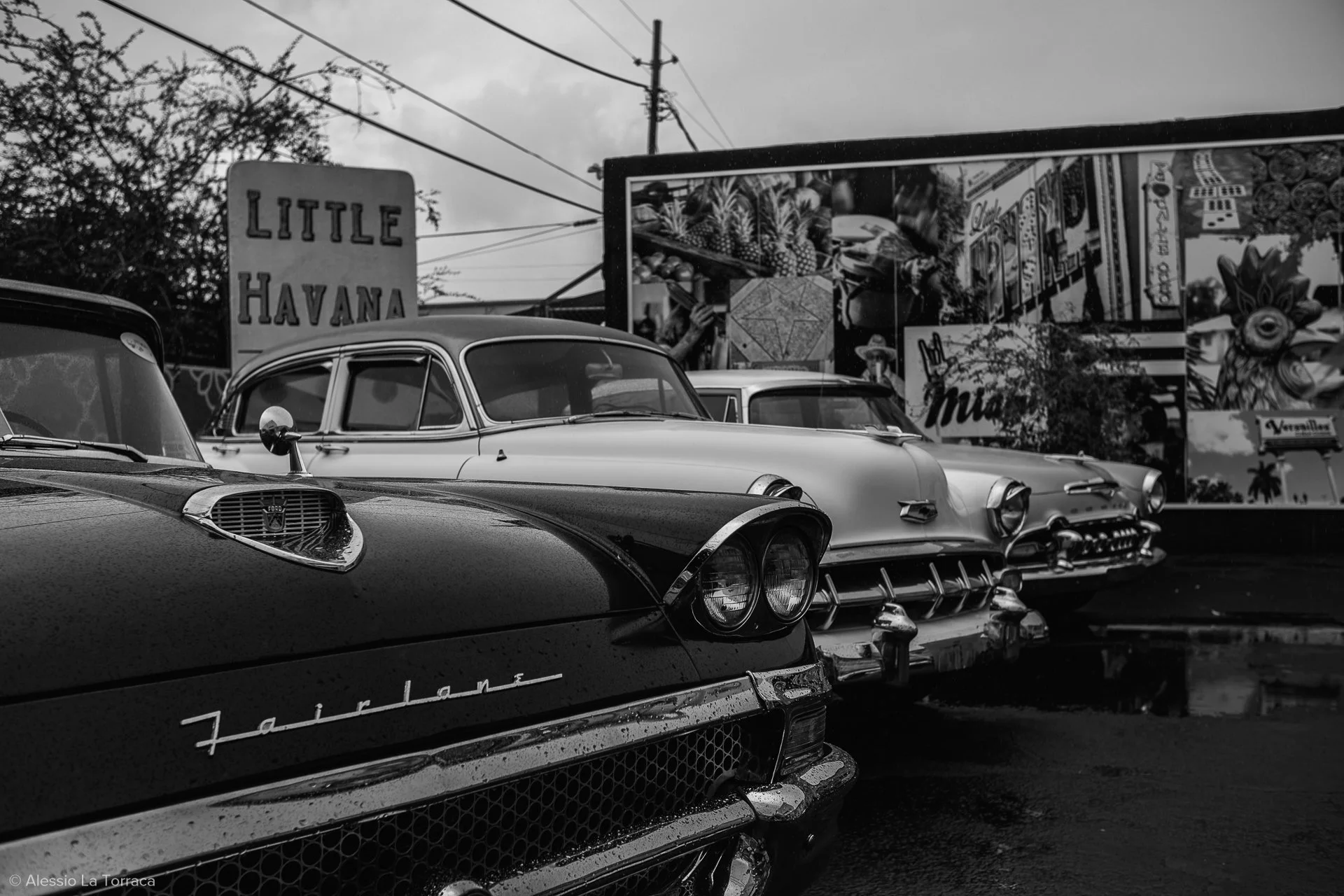 Black and white photo of vintage cars parked in a row in front of a sign that says "Little Havana" with a billboard featuring colorful images of pineapples, a taco, and other vibrant items, indicating a lively, possibly Cuban-themed area.