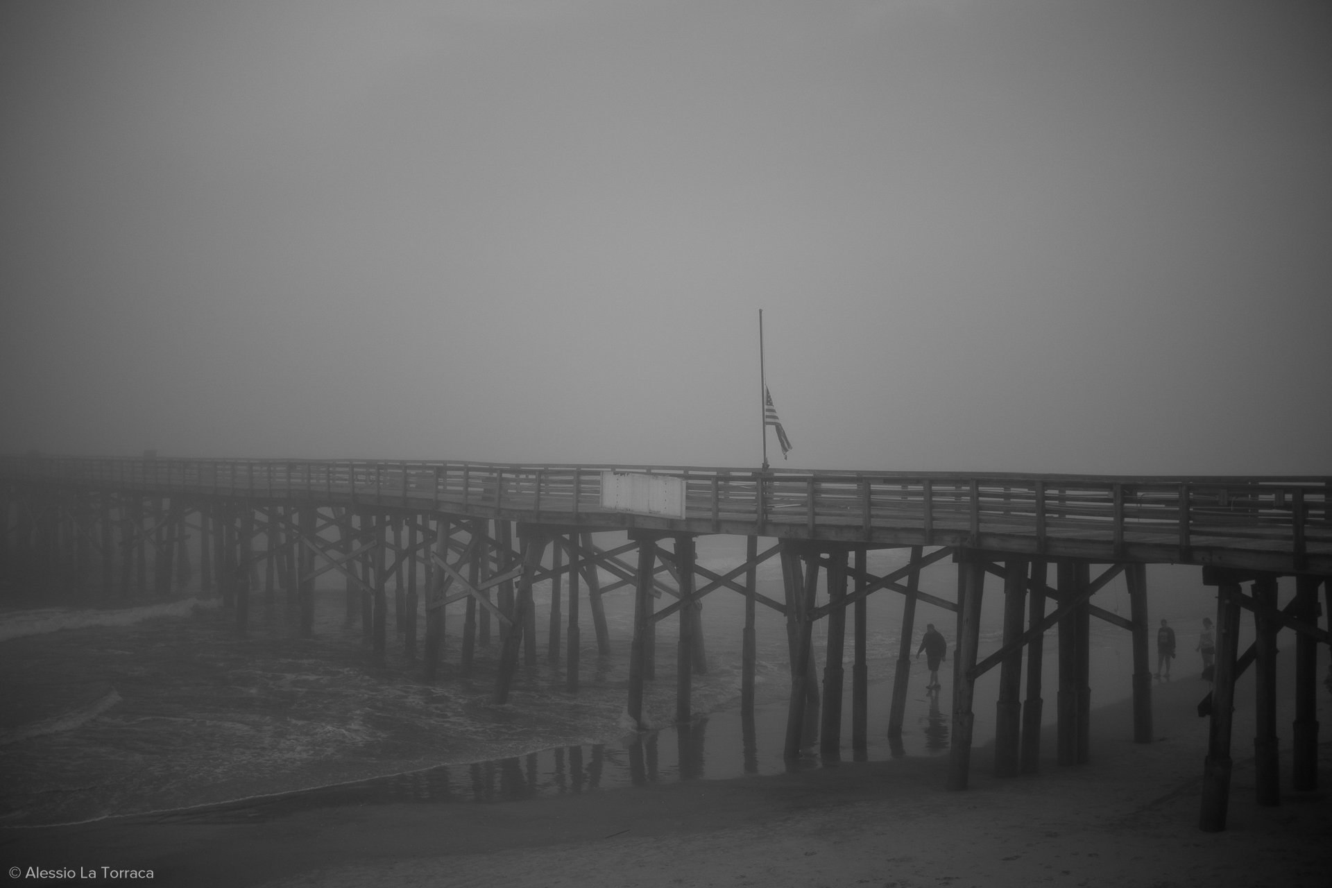 A foggy scene of a wooden pier extending over the ocean with an American flag flying at the end, and a few people walking along the shore beneath it.