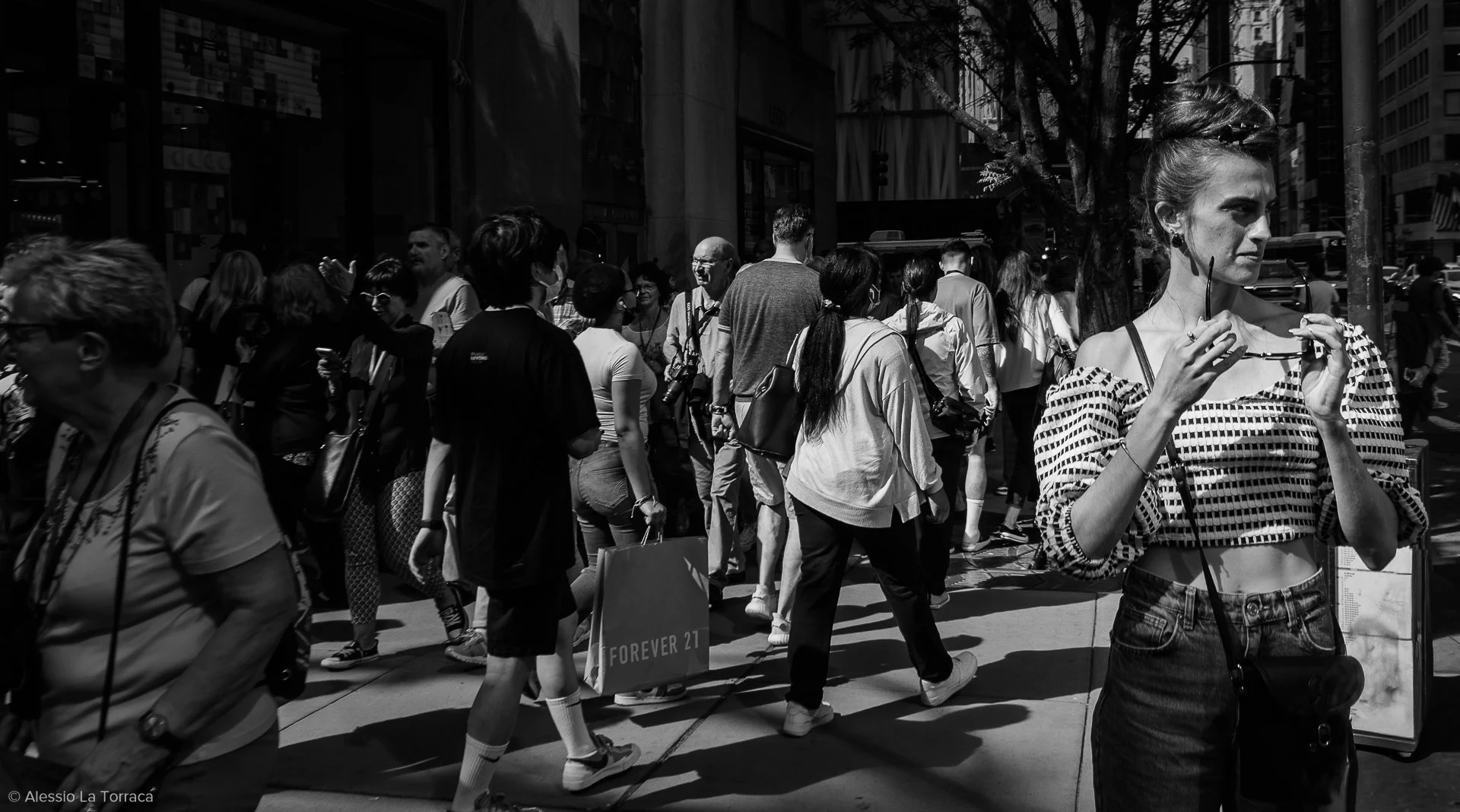 A busy city sidewalk filled with crowd of people walking and standing, with buildings and trees in the background. A woman in the foreground holds sunglasses near her face.
