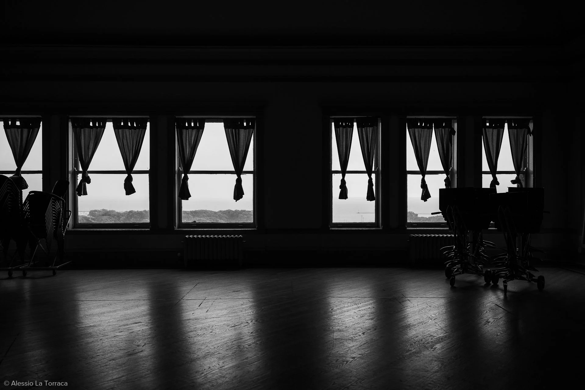 Silhouetted interior of a room with large windows and curtains, showing a landscape outside, with stacked chairs and shopping carts inside.