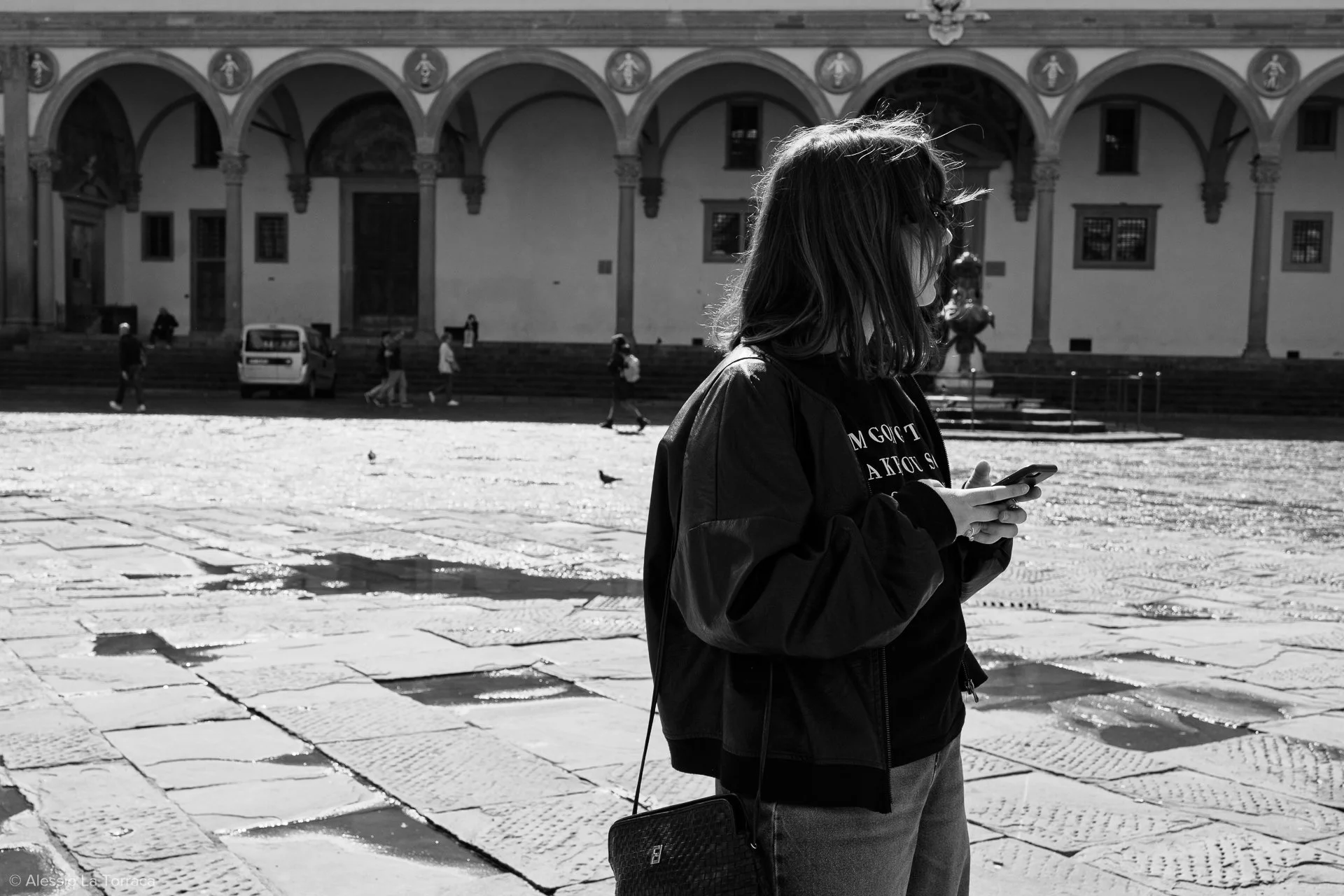 A young woman with shoulder-length hair looking at her phone on a sunny cobblestone street with a historic building in the background.
