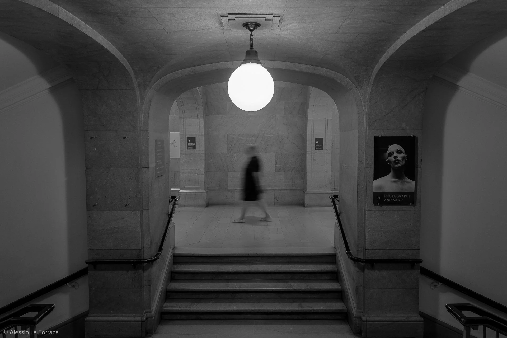 A person walking through an art gallery or museum with marble walls and stairs, illuminated by a spherical ceiling light, with a portrait photograph on the right wall.