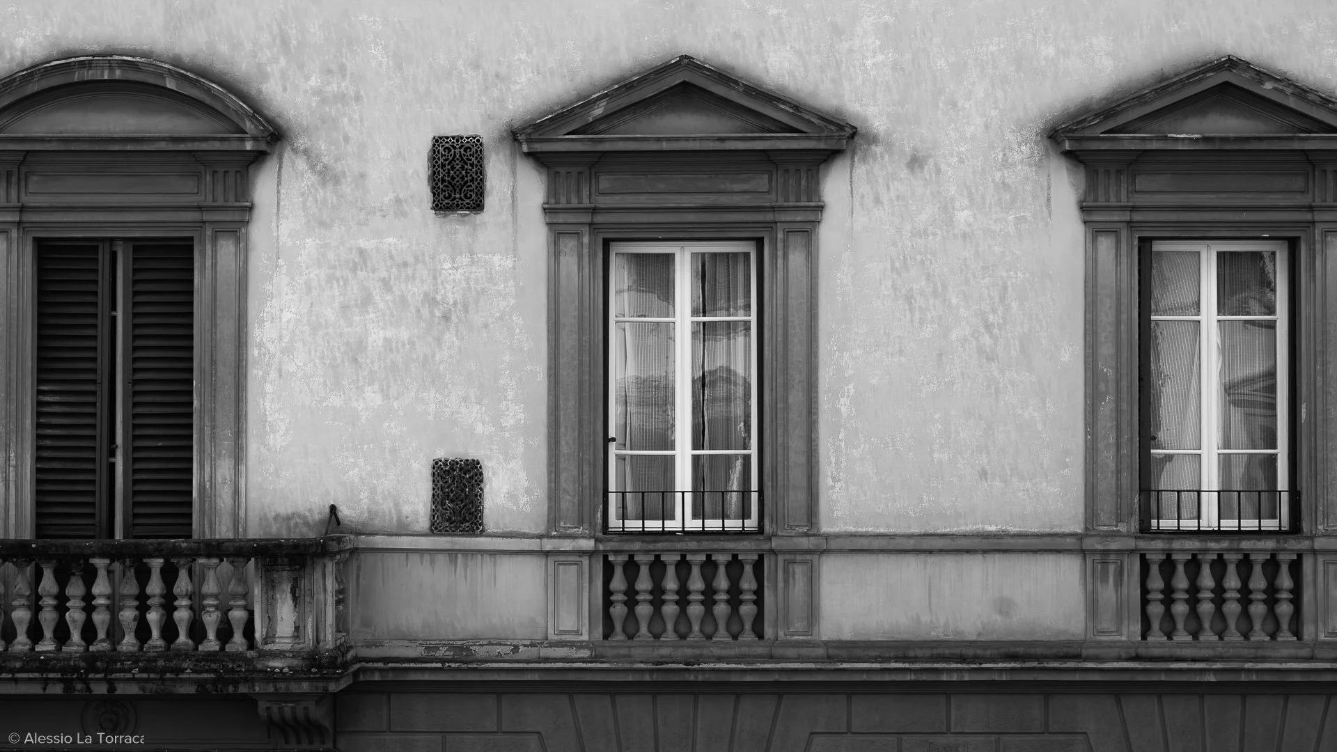 Black and white photo of a building's facade with three windows; the middle window has glass doors with a small balcony, and the windows have decorative wooden trim and shutters.