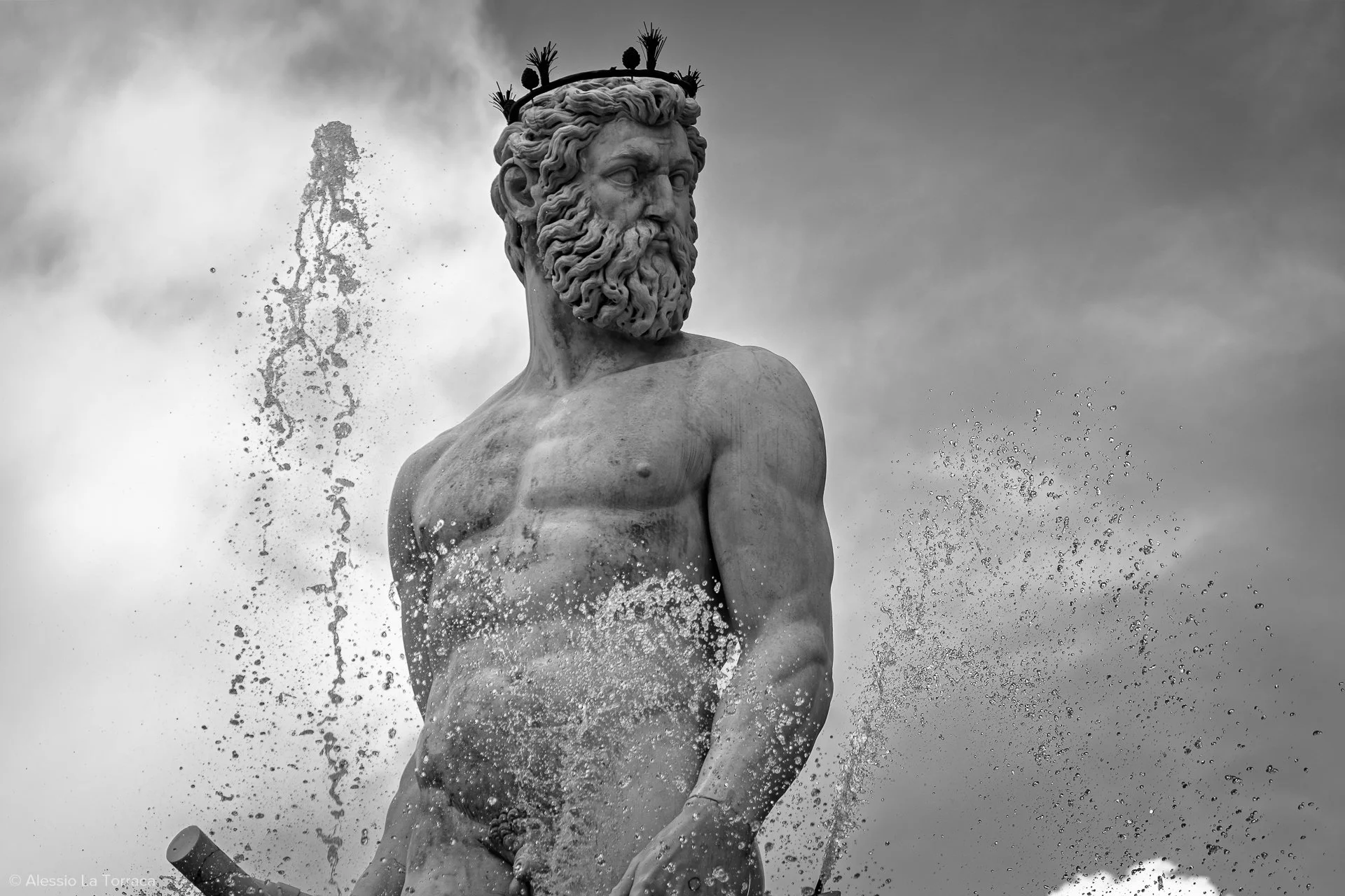 Black and white photograph of a classical stone statue of a bearded man with a crown of plants, partially submerged in water with water splashing around.