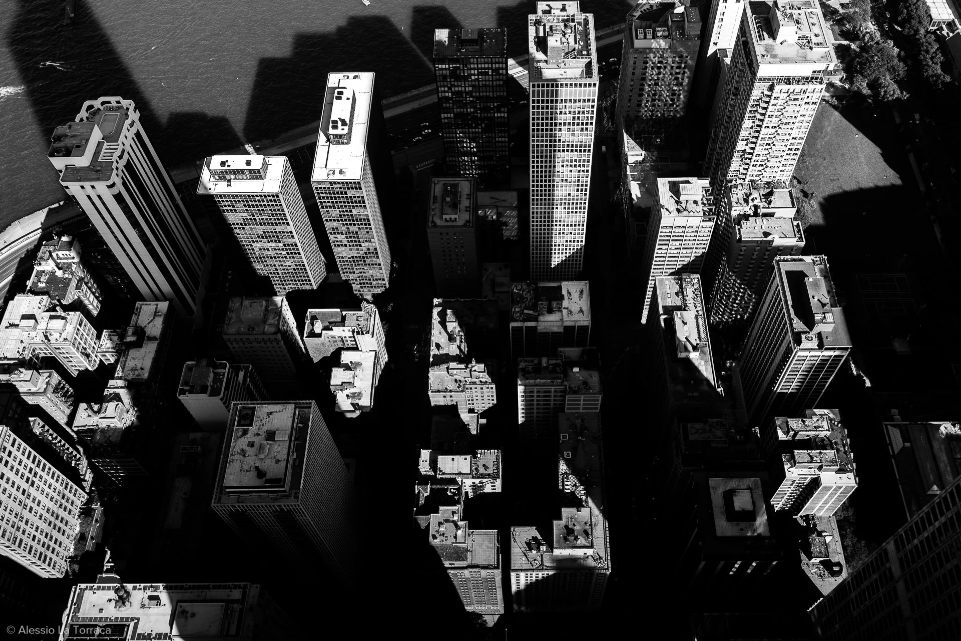 Black and white aerial view of a cityscape with tall skyscrapers blocking sunlight, casting shadows, and a river running alongside buildings.