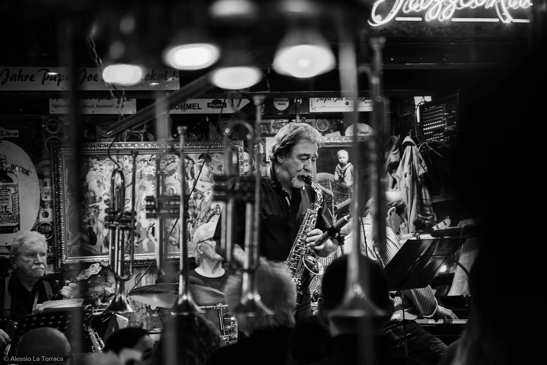 A black and white photo of a live music performance featuring a saxophonist, a drummer, and a pianist, with a crowd in the foreground and colorful artwork on the walls behind the musicians.