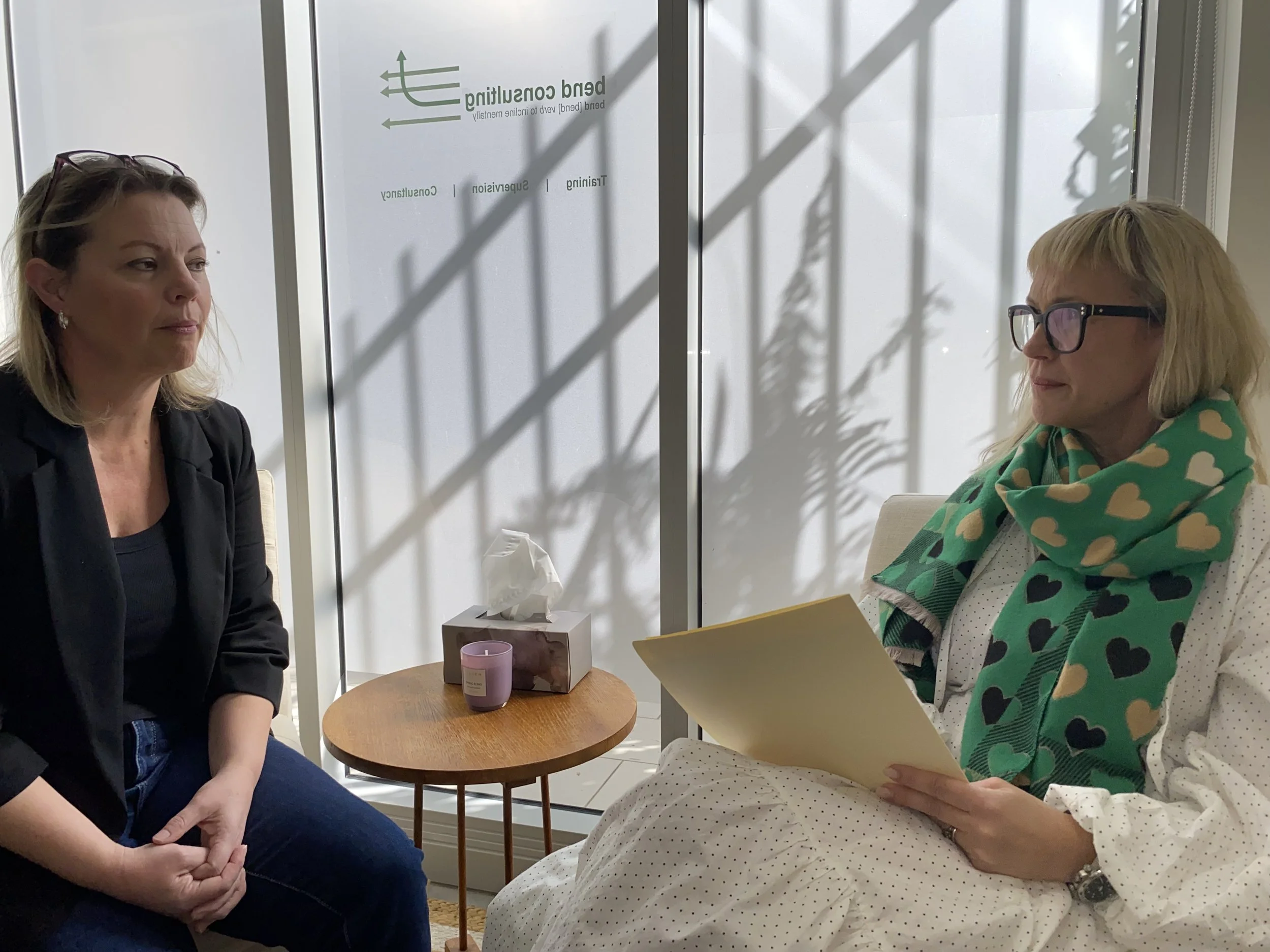 Two women sitting in a consultation room, engaged in conversation. One woman is wearing glasses and a green scarf with heart patterns, holding a yellow folder. The other woman has blonde hair and is dressed in a black blazer. There is a small round wooden table with a tissue box and a purple candle between them, with a glass door behind them casting shadows of plant leaves.