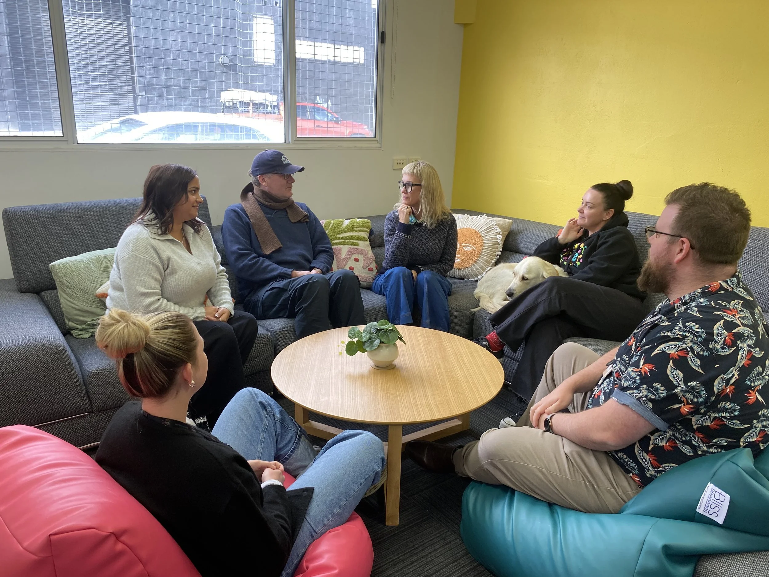 Six people and two dogs sitting in a semi-circle in a room with yellow and white walls, engaging in a conversation around a round wooden table with a small potted plant. There are windows in the background showing parked cars.
