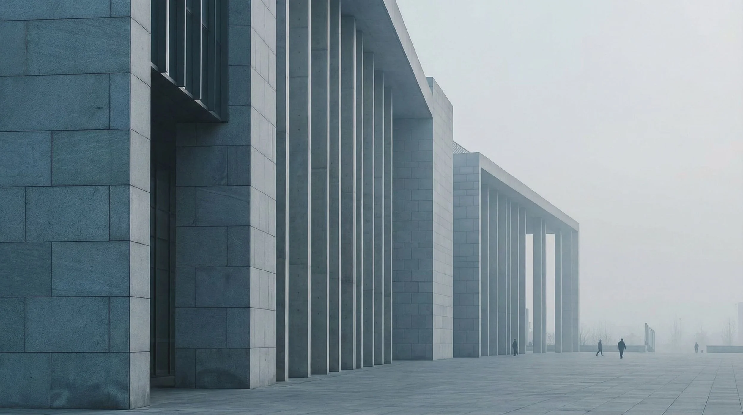 Wide view of a modern stone public building with tall columns, partly obscured by fog, with a few people on a plaza in the distance.