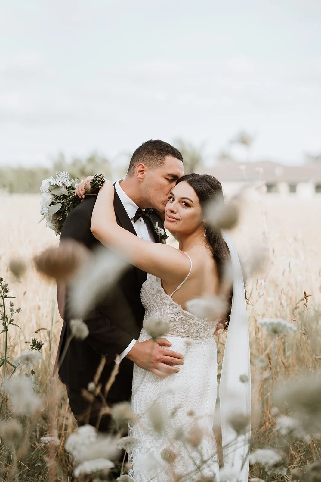 A newlywed couple embracing in a field, with the groom kissing the bride's temple and the bride smiling at the camera, surrounded by tall grass and flowers.