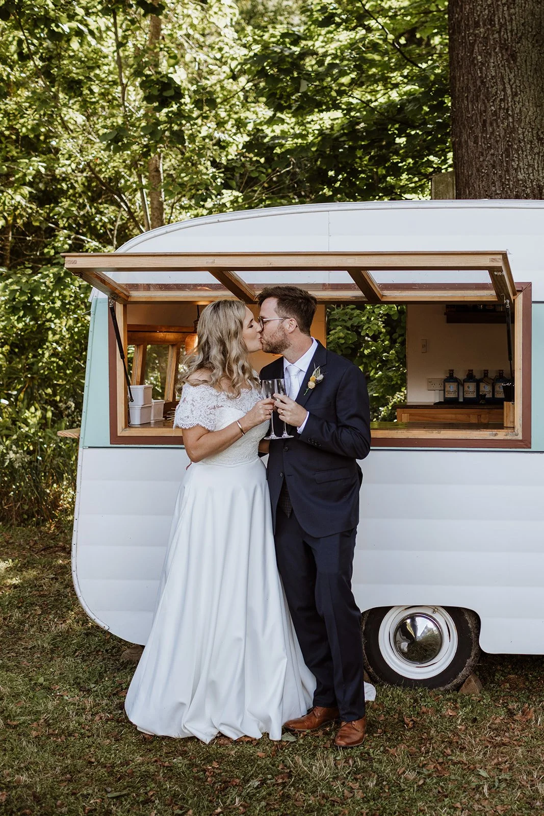 A bride and groom sharing a kiss and holding champagne glasses in front of a vintage trailer at an outdoor wedding.