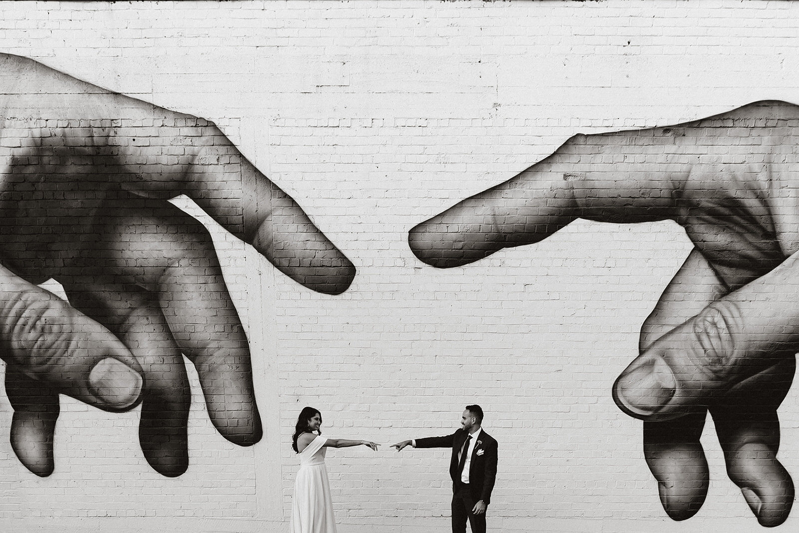 A woman and a man in formal attire holding hands in front of a large black-and-white mural of two hands reaching towards each other on a brick wall.