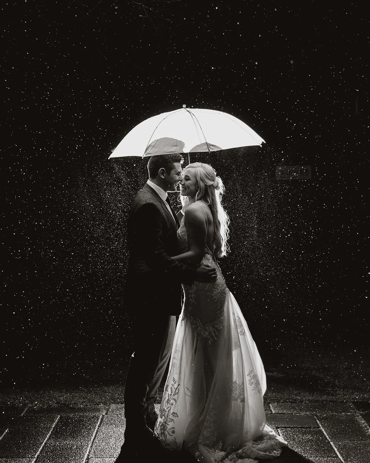 A black and white photo of a couple in wedding attire holding under an umbrella in the rain, embracing and smiling at each other, with rain droplets falling around them.