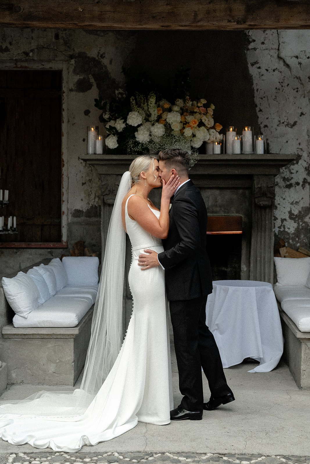 A bride and groom sharing a kiss during their wedding ceremony, with the bride in a white dress and veil, and the groom in a black suit, in front of a rustic stone fireplace with candles and floral arrangement.