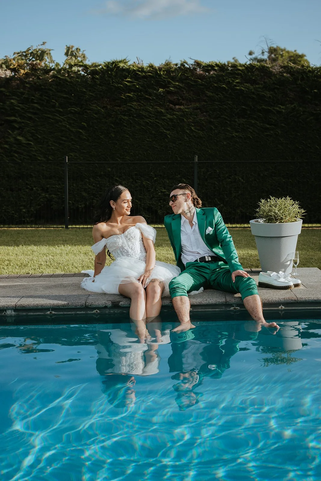 A woman in a white dress and a man in a green suit sit by a swimming pool, with their feet immersed in the water, smiling and looking at each other. The background features a tall hedge and clear blue sky.