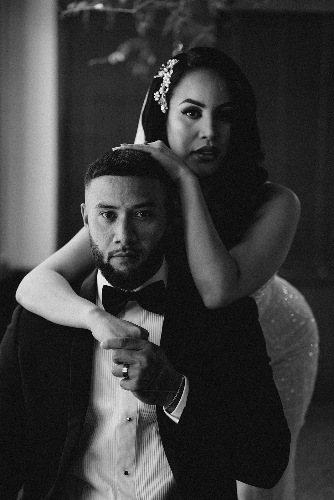 A black and white photo of a elegant couple, with the man in a tuxedo and the woman with dark hair and a decorative hair accessory, posing closely together