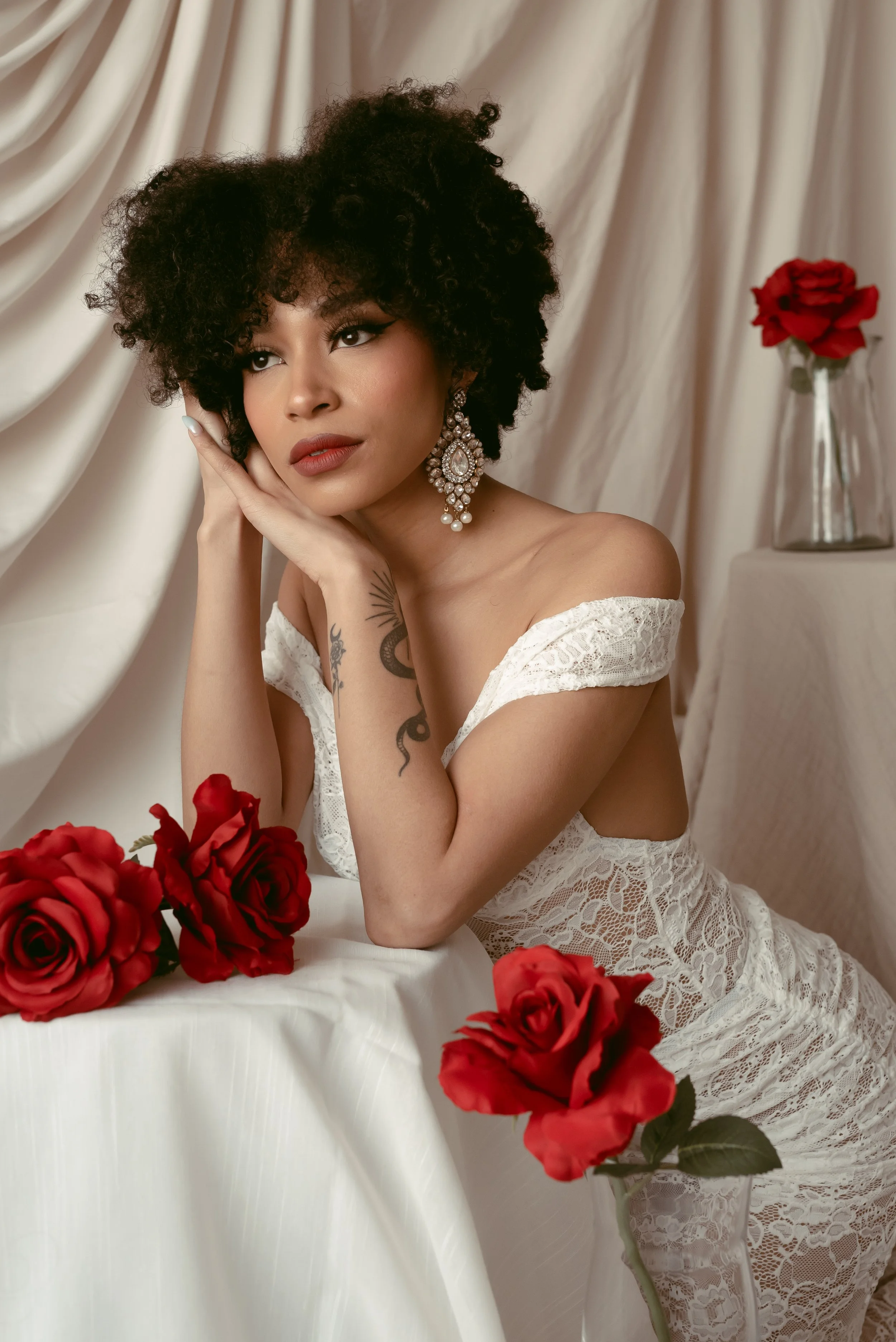 A woman with curly dark hair and earrings, wearing a white lace dress, resting her head on her hand amidst red roses, standing near a table with a cream-colored cloth, in front of cream drapes.