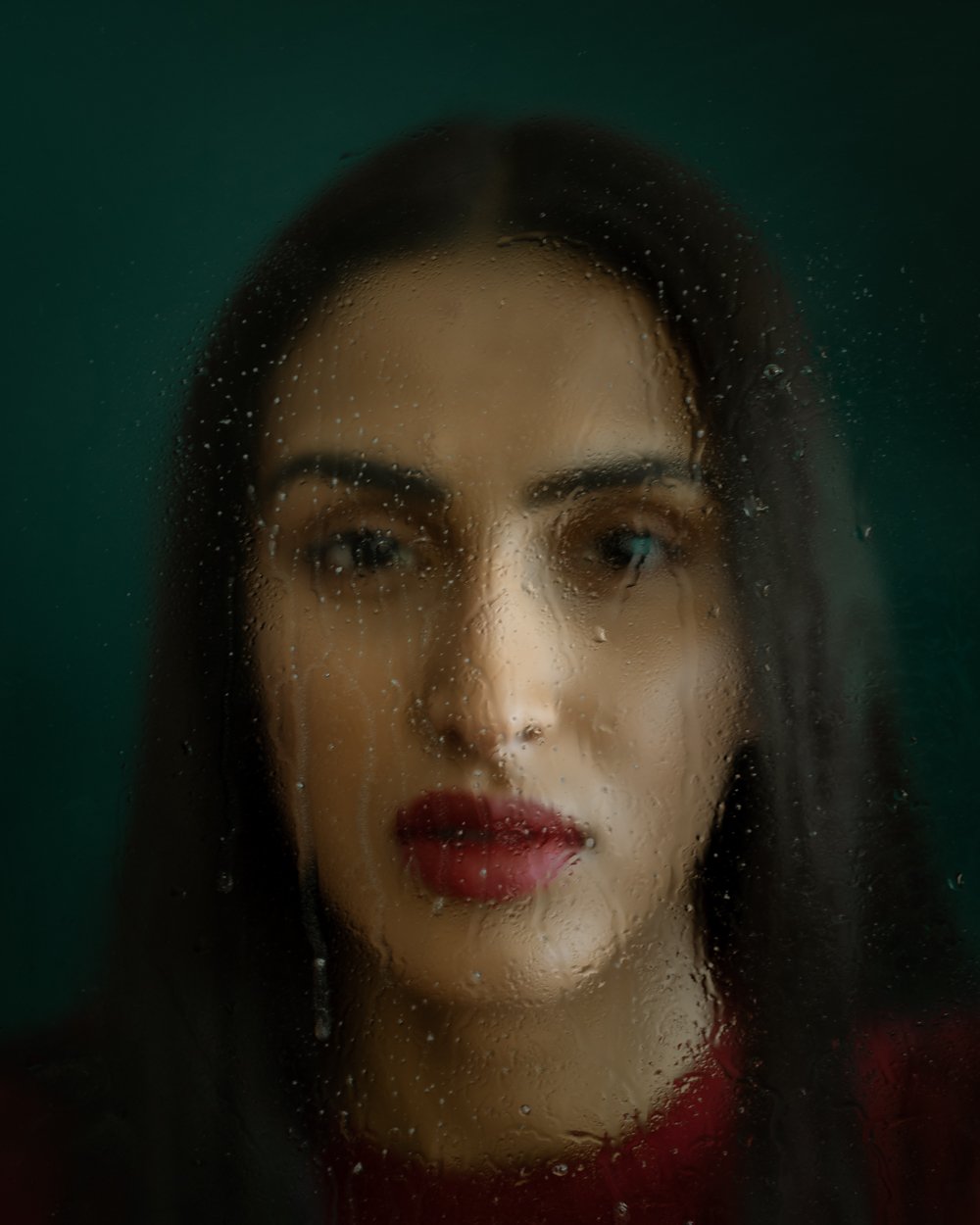 Portrait of a woman with dark hair and red lipstick, viewed through a foggy glass with water droplets.