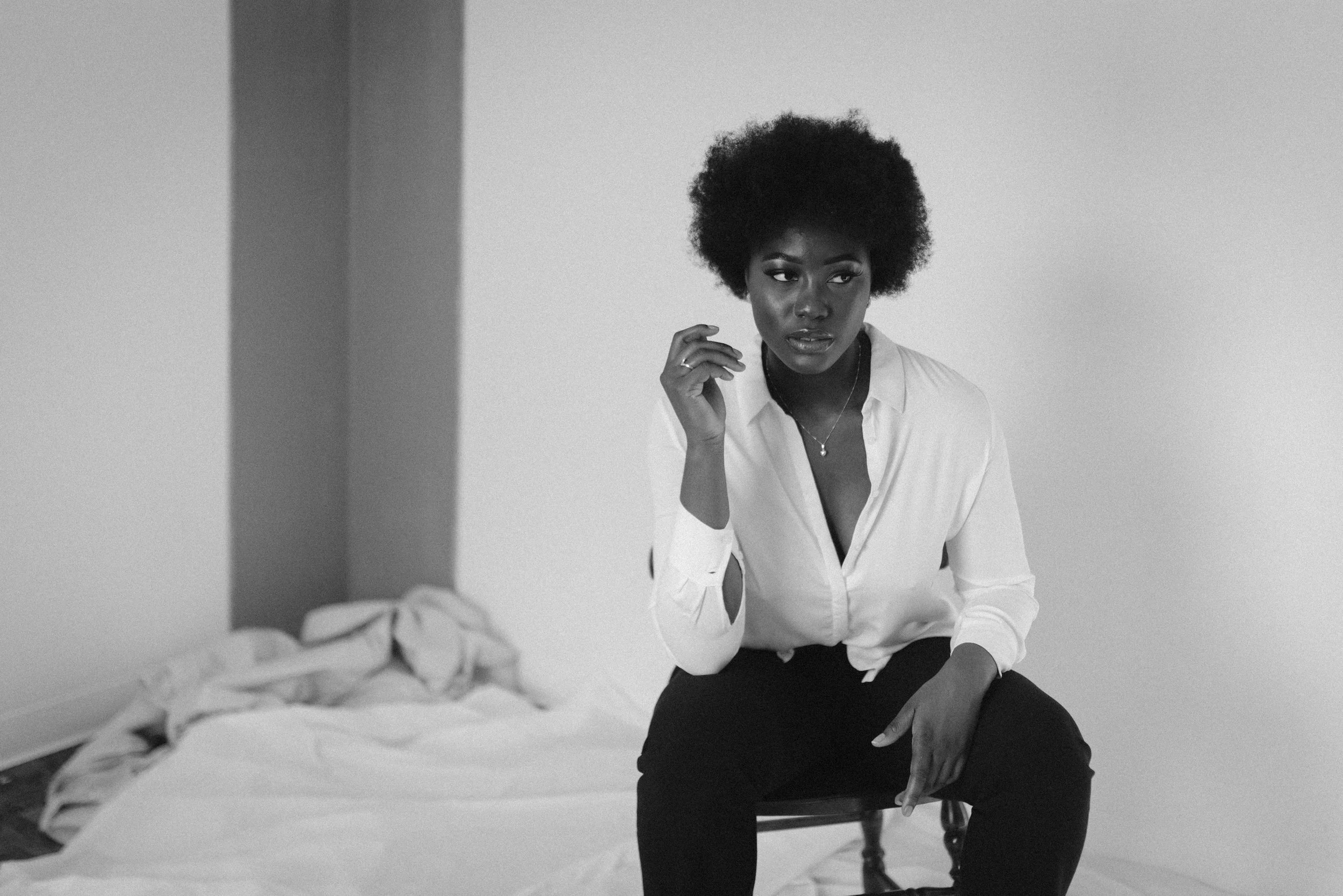 A woman with a large afro hairstyle wearing a white shirt sitting on a chair in a room with plain walls and a bed with rumpled sheets in the background.