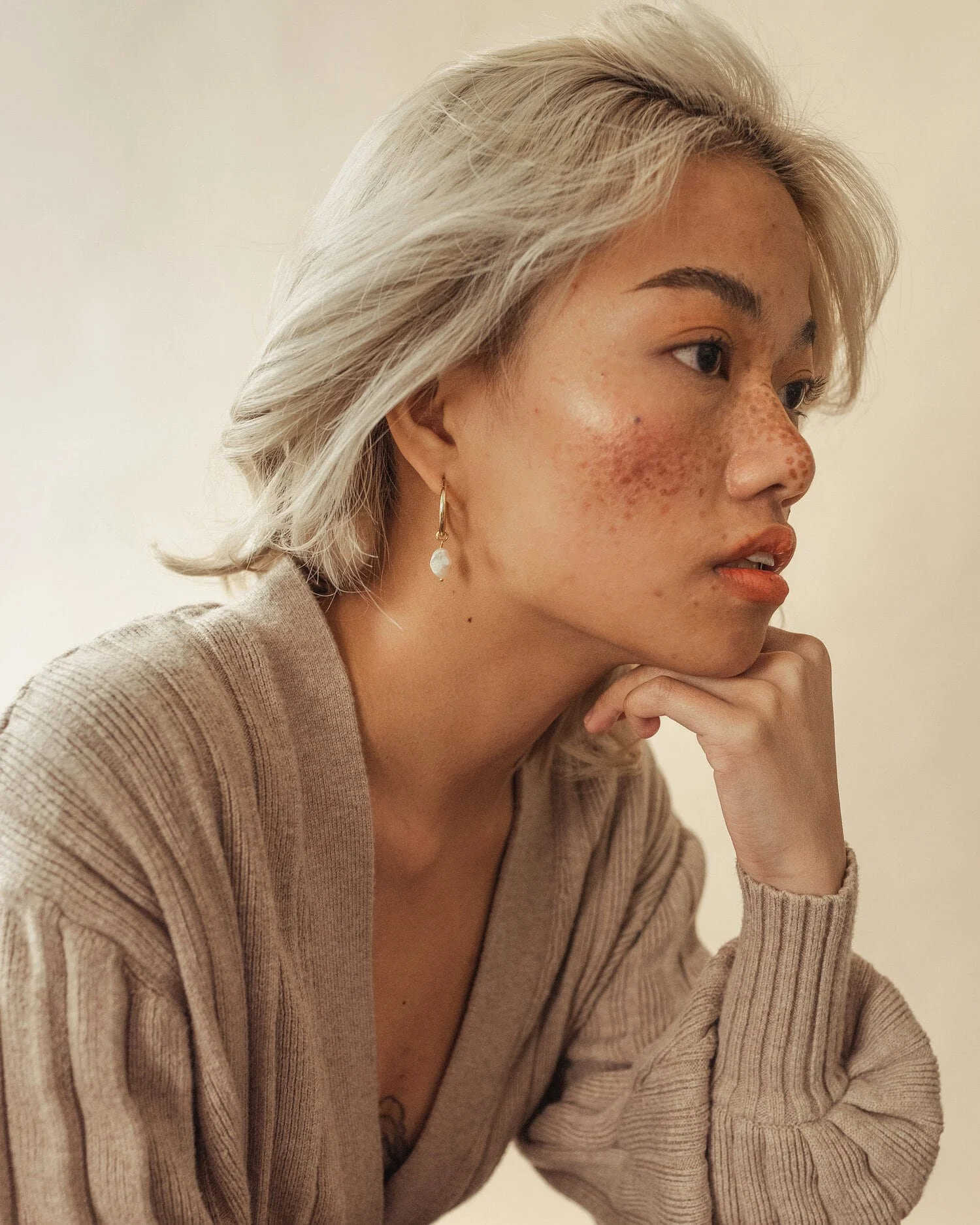A woman with blonde hair, wearing a beige sweater and a dangling earring, resting her chin on her hand, gazing thoughtfully to the side.