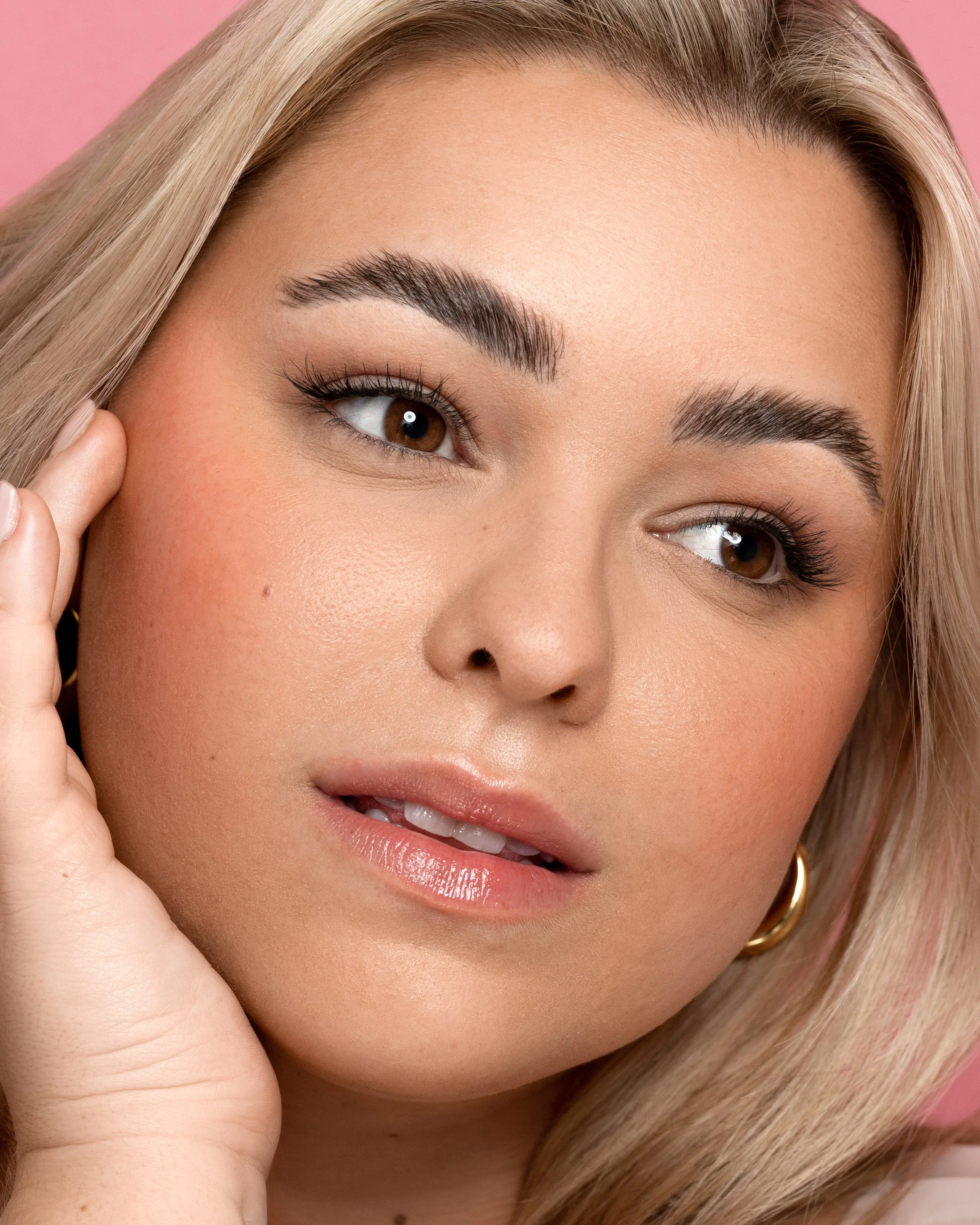 Close-up of a young woman's face with blonde hair, brown eyes, and makeup, touching her face gently with her left hand.