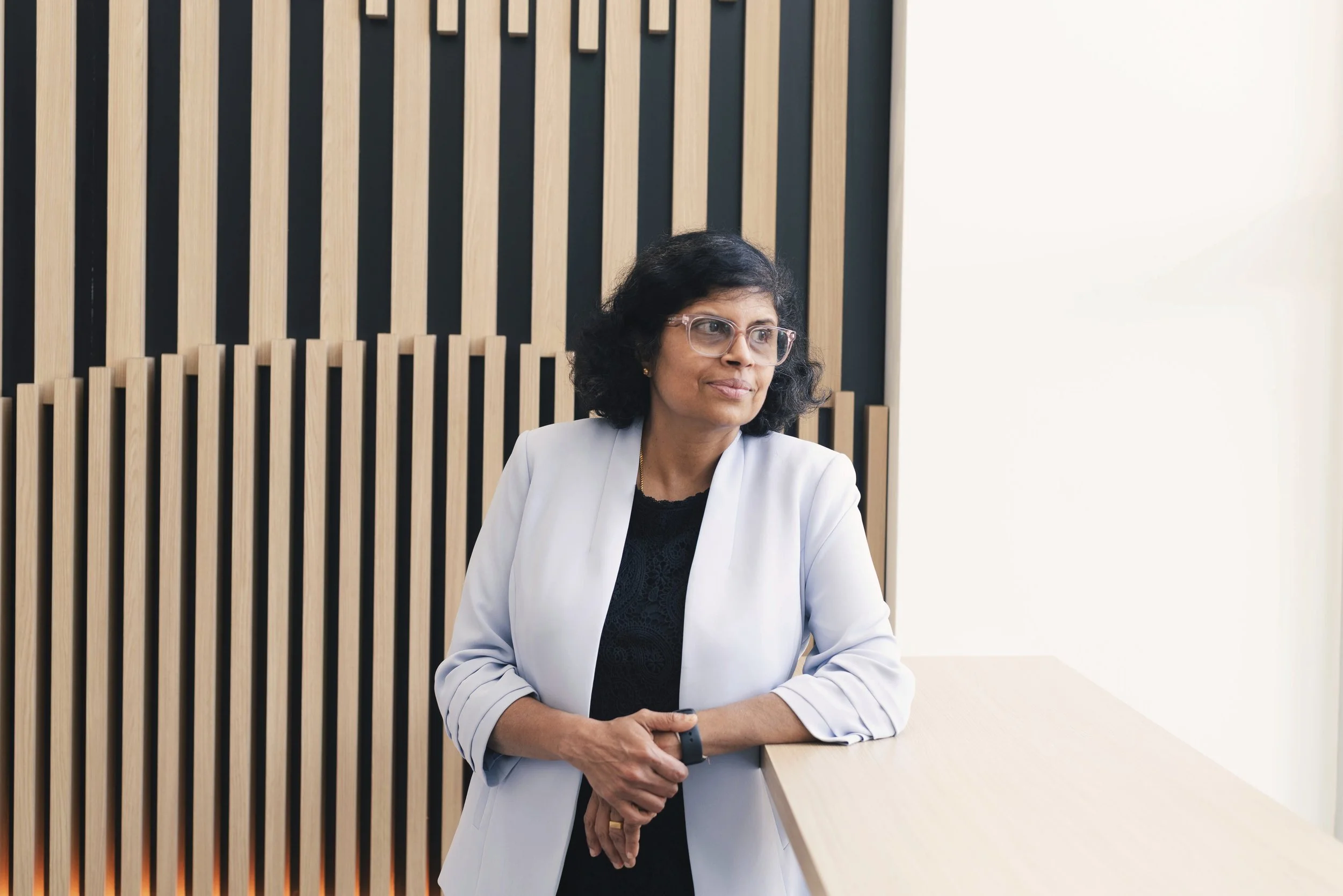 A woman with glasses and dark curly hair standing at a counter, wearing a white blazer and black top, with a wooden slat background and bright lighting.