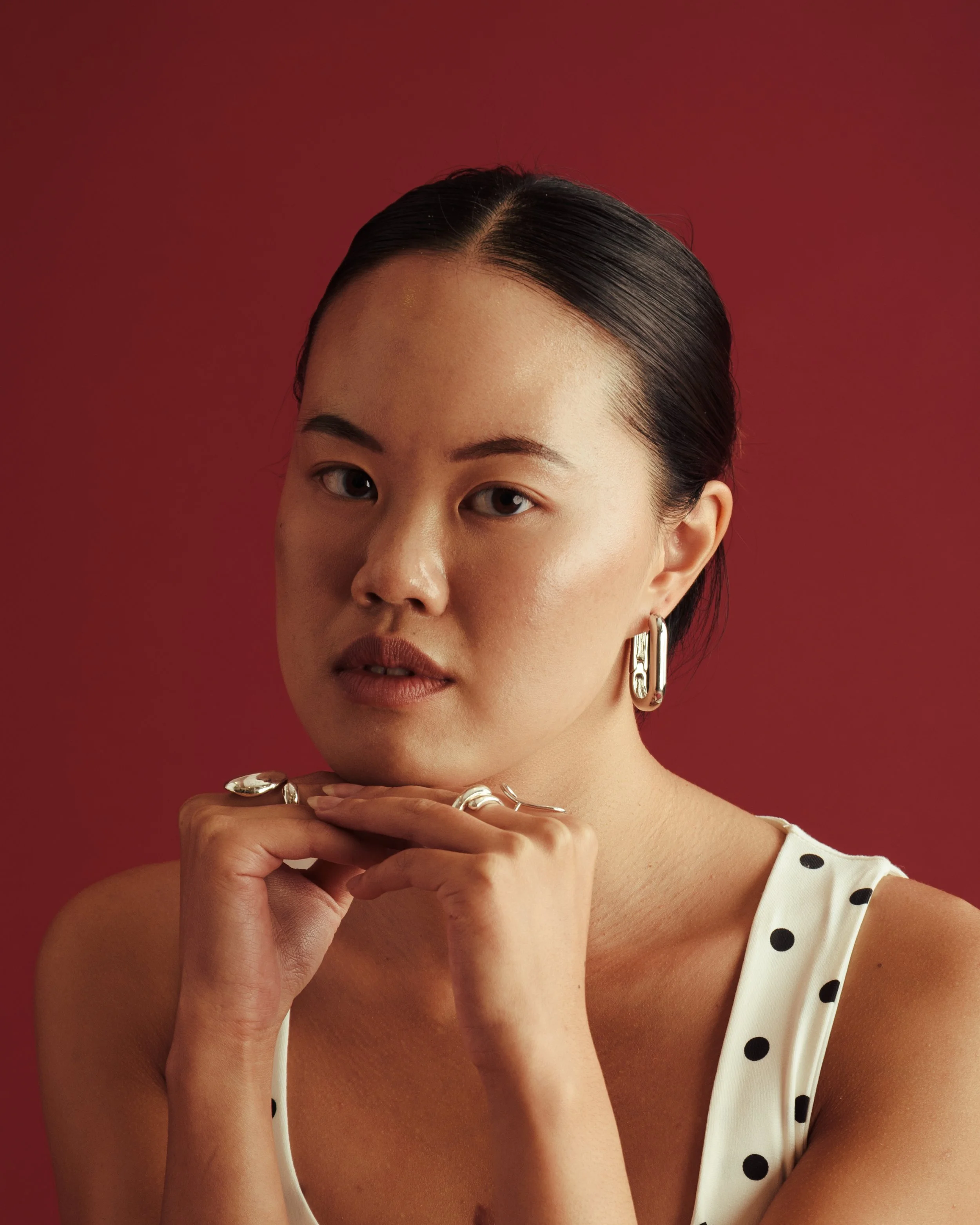 A woman with dark hair slicked back, wearing silver jewelry and a white dress with black polka dots, posing against a red background.