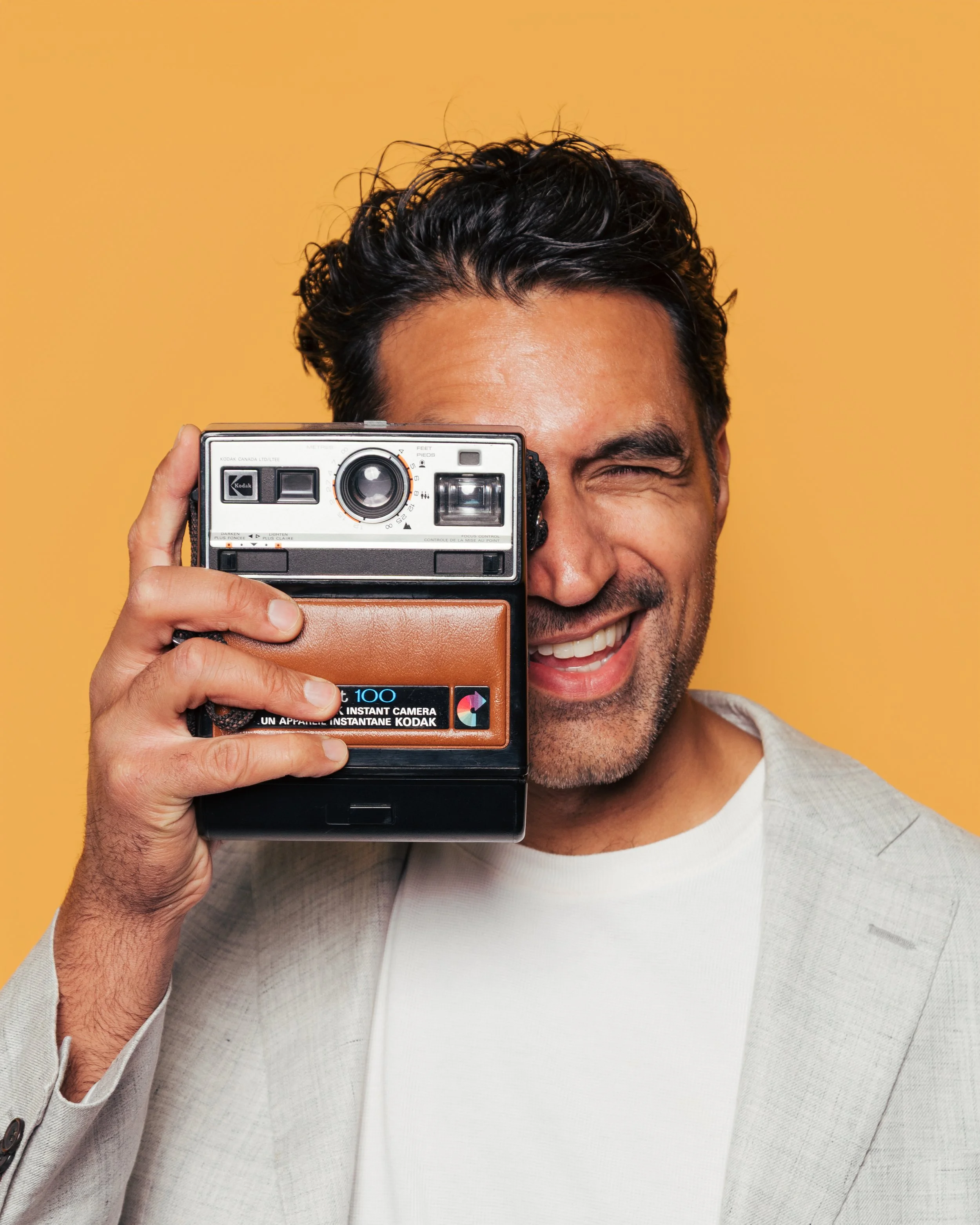 A man with dark hair and a light beard, smiling and squinting, holding a vintage camera up to his eye against a yellow background, wearing a light-colored jacket and white shirt.