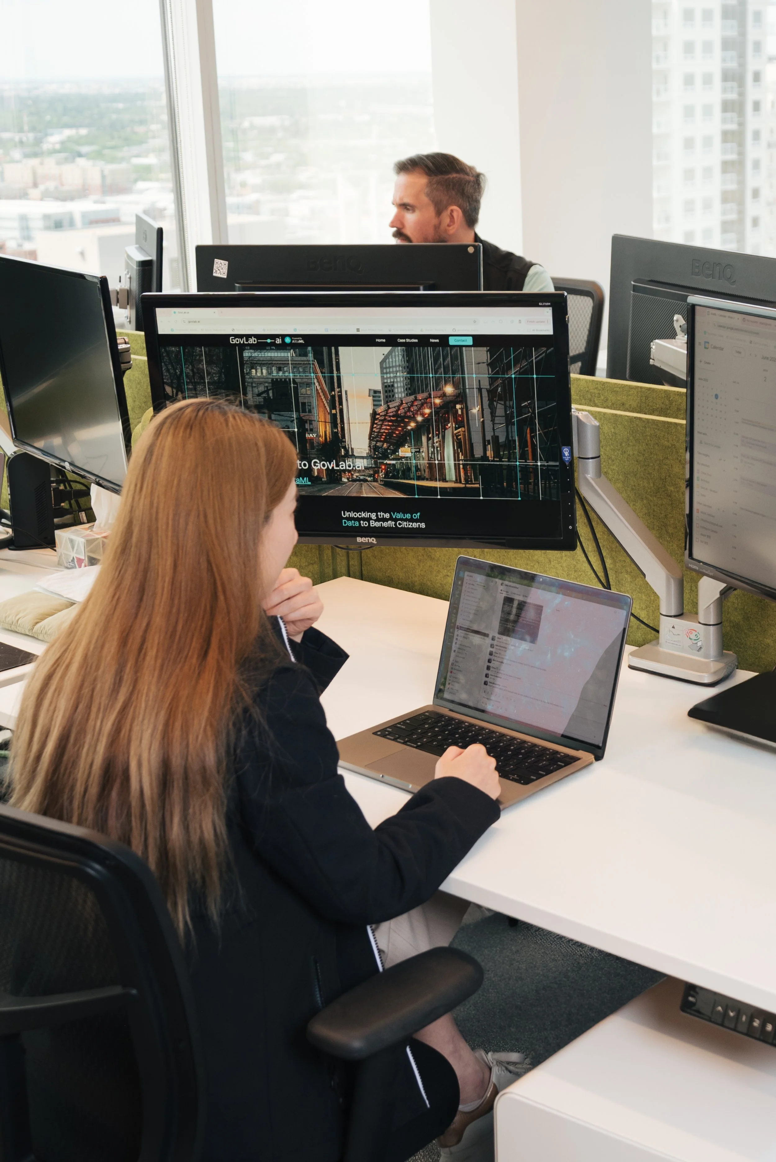 A woman with long red hair working at her desk in an office, with multiple monitors and a laptop, and a man working at his desk in the background near large windows with a city view.