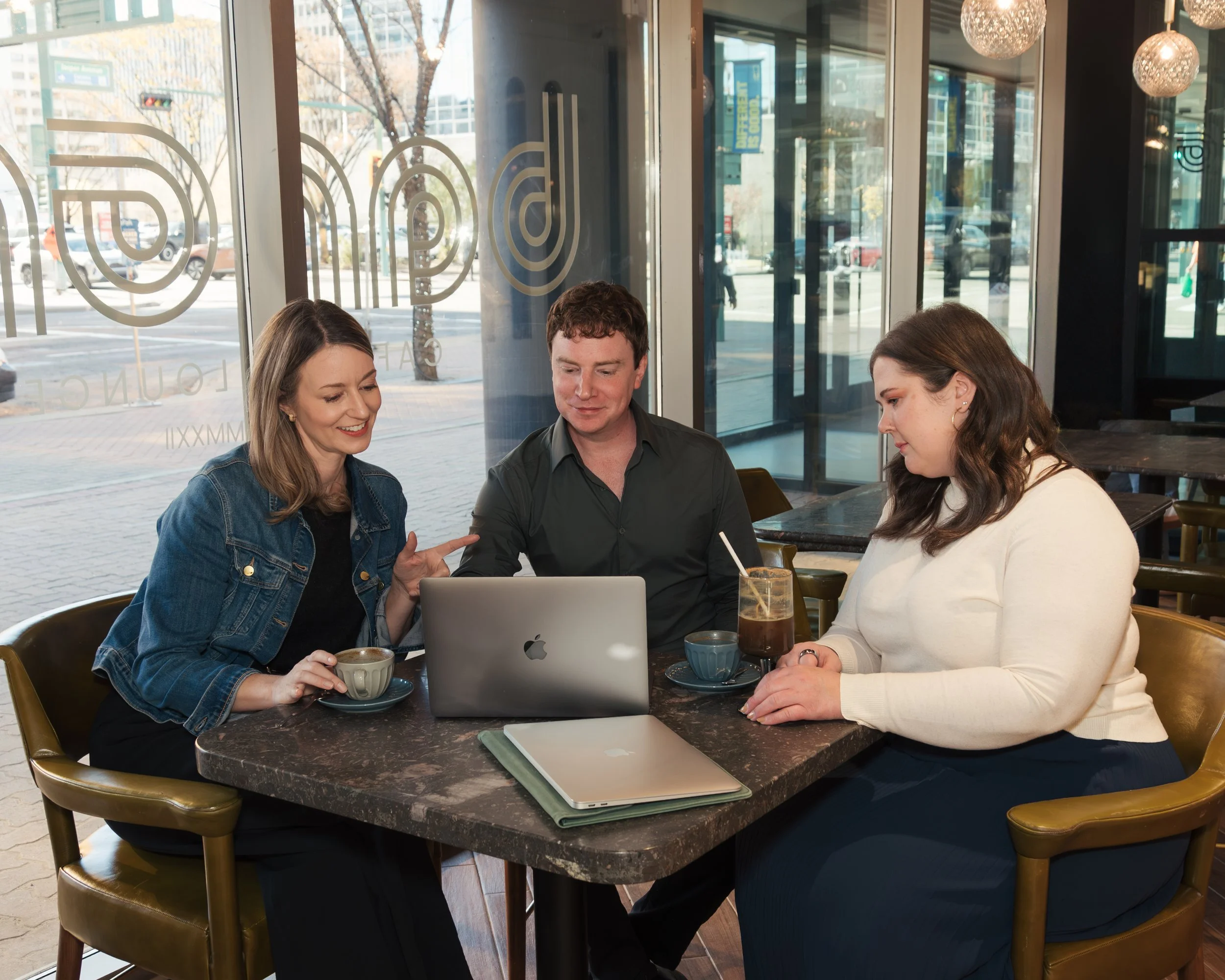 Three friends sitting at a table in a coffee shop, looking at a laptop, with drinks, engaging in conversation.