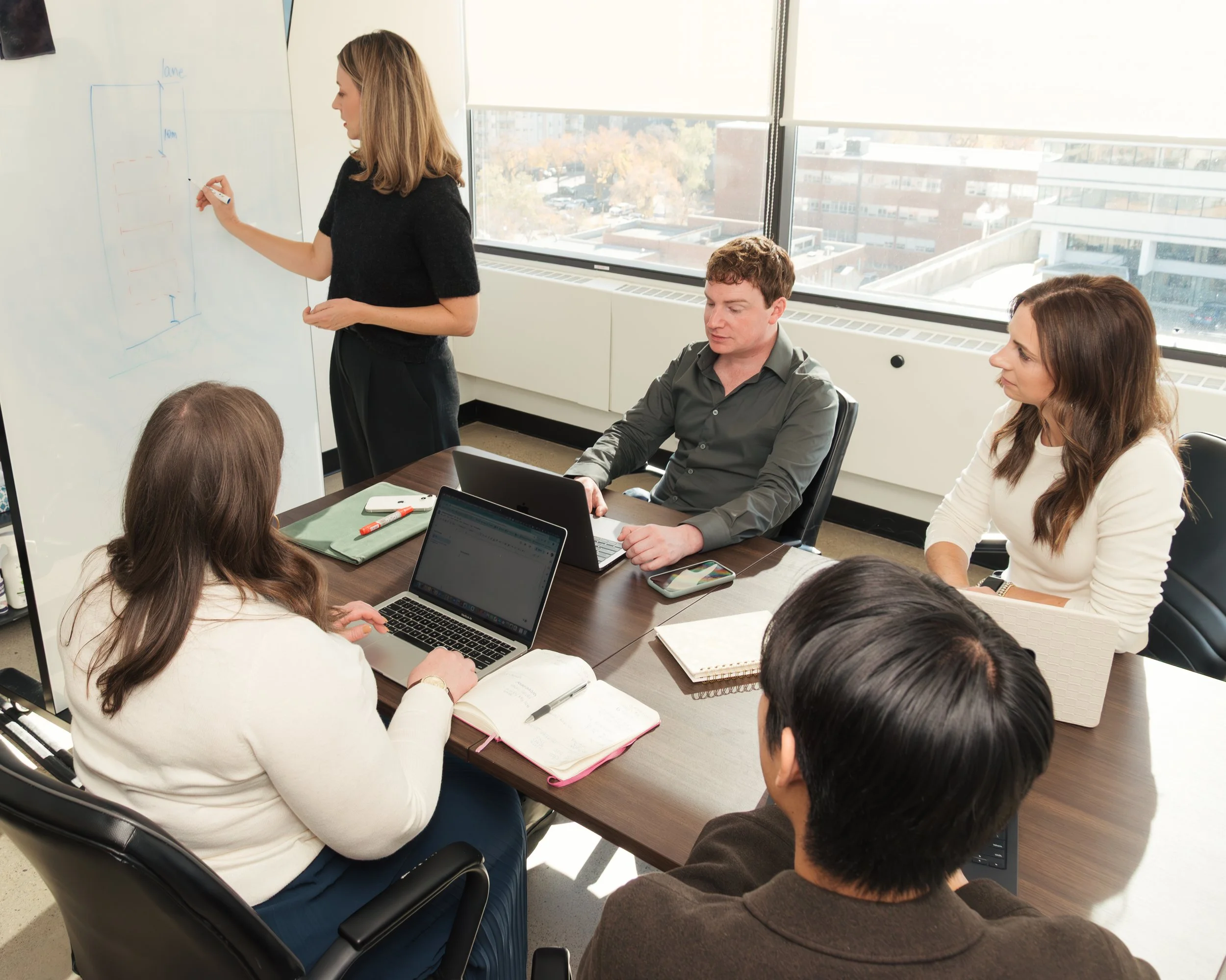A woman stands at a whiteboard drawing diagrams while giving a presentation to five colleagues seated around a conference table. The colleagues are working on laptops and taking notes as they listen.