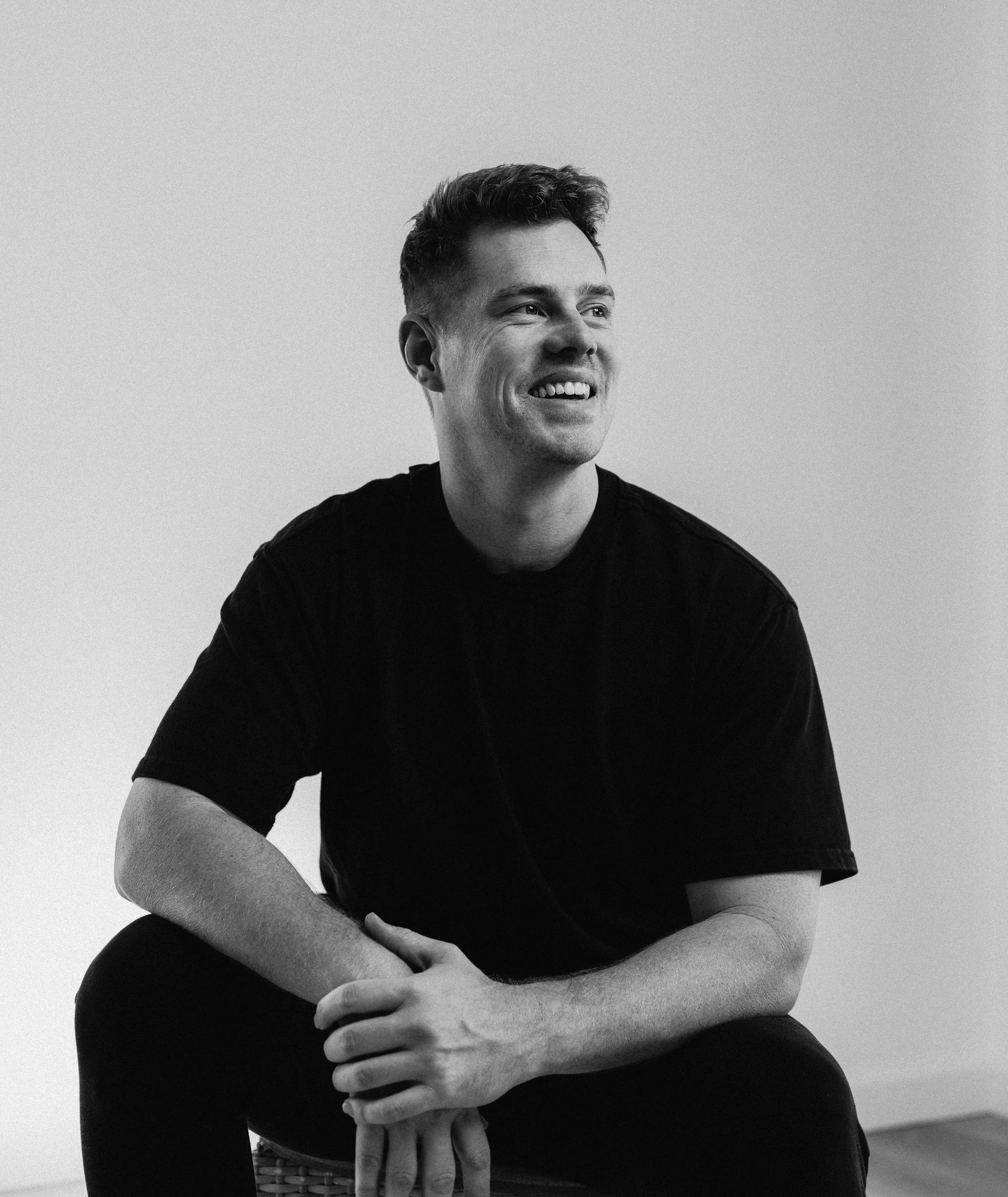 Black and white photograph of a young man with short hair smiling, sitting with his arms crossed over his knees, wearing a black t-shirt, against a plain light background.