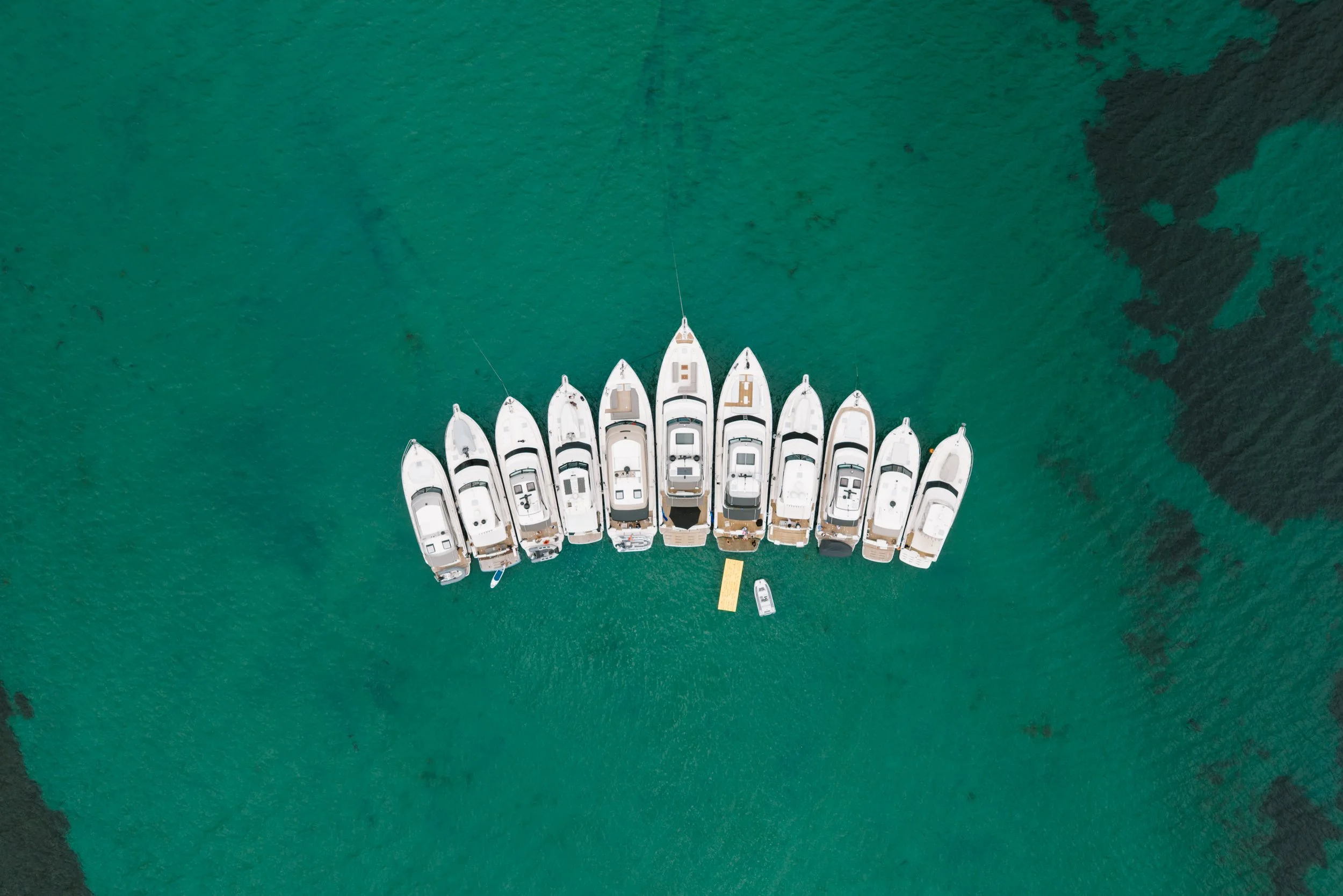 Top-down view of several white yachts docked closely together in a marina, surrounded by clear greenish water.