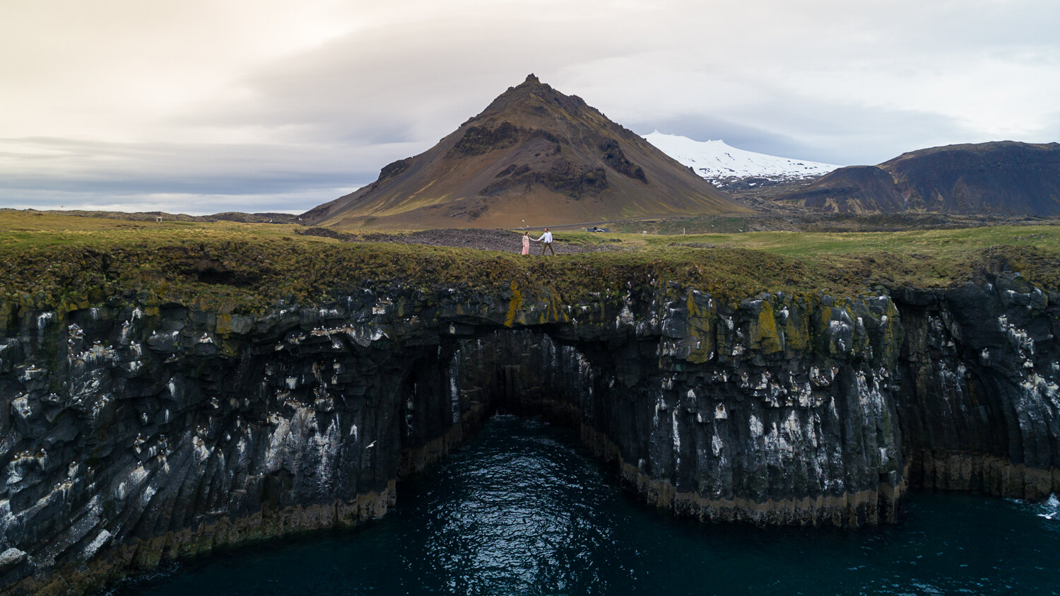 Iceland elopement photographer. Drone elopement photography of a couple on rock arch bridge over ocean at Arnarstapi in Iceland.