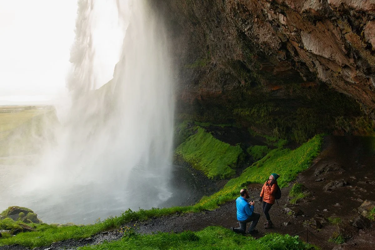 Proposal behind Seljalandsfoss waterfall in Iceland. He is proposing and she is surprised and emotional. Photo by Iceland Proposal Photographer.