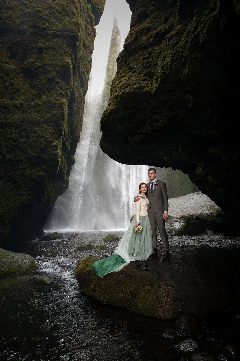 Engagement photo session inside a gorge with a hidden waterfall in Iceland. Couple is wearing nice attire. Green dress skirt and grey suit with blue tie.