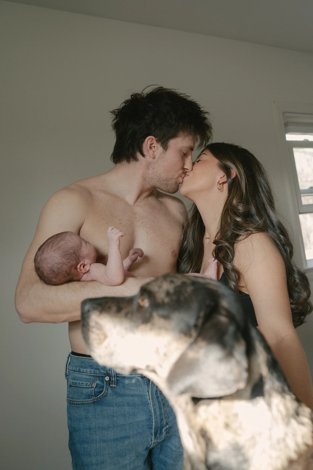 Parents kissing in a family photoshoot with their newborn baby and dog