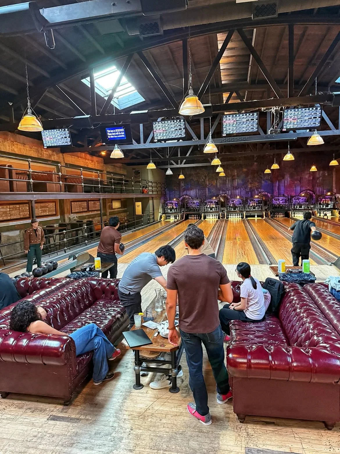 A group of people bowling, relaxing, and having food on leather couches inside Highland Park Bowl.