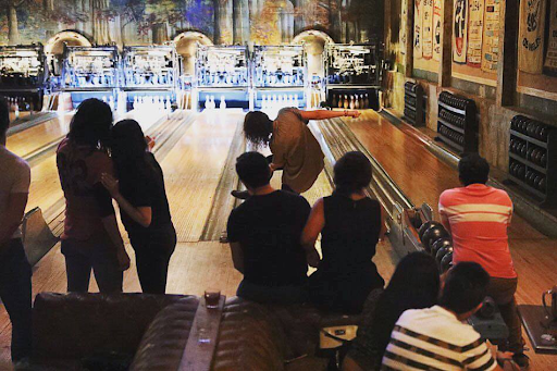 Bowler rolling a ball down a lane while friends watch from lounge seating at Highland Park Bowl.
