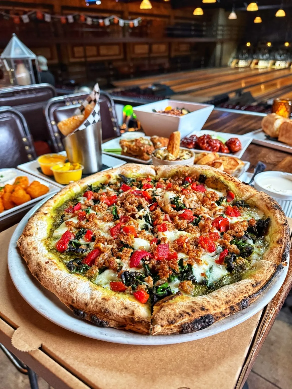 Wood-fired pizza, chicken wings, and shareable appetizers on a dining table overlooking the bowling lanes at Highland Park Bowl.