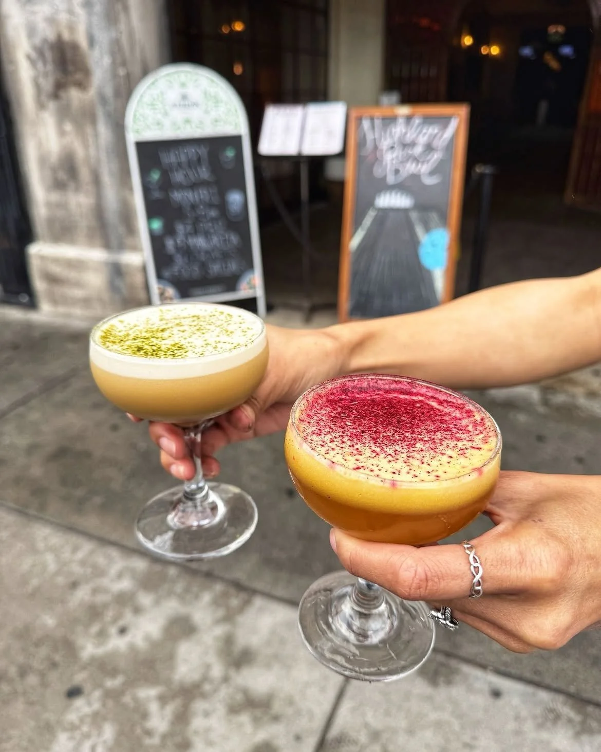 Two hands raising colorful craft cocktails in front of the illuminated Highland Park Bowl sign.